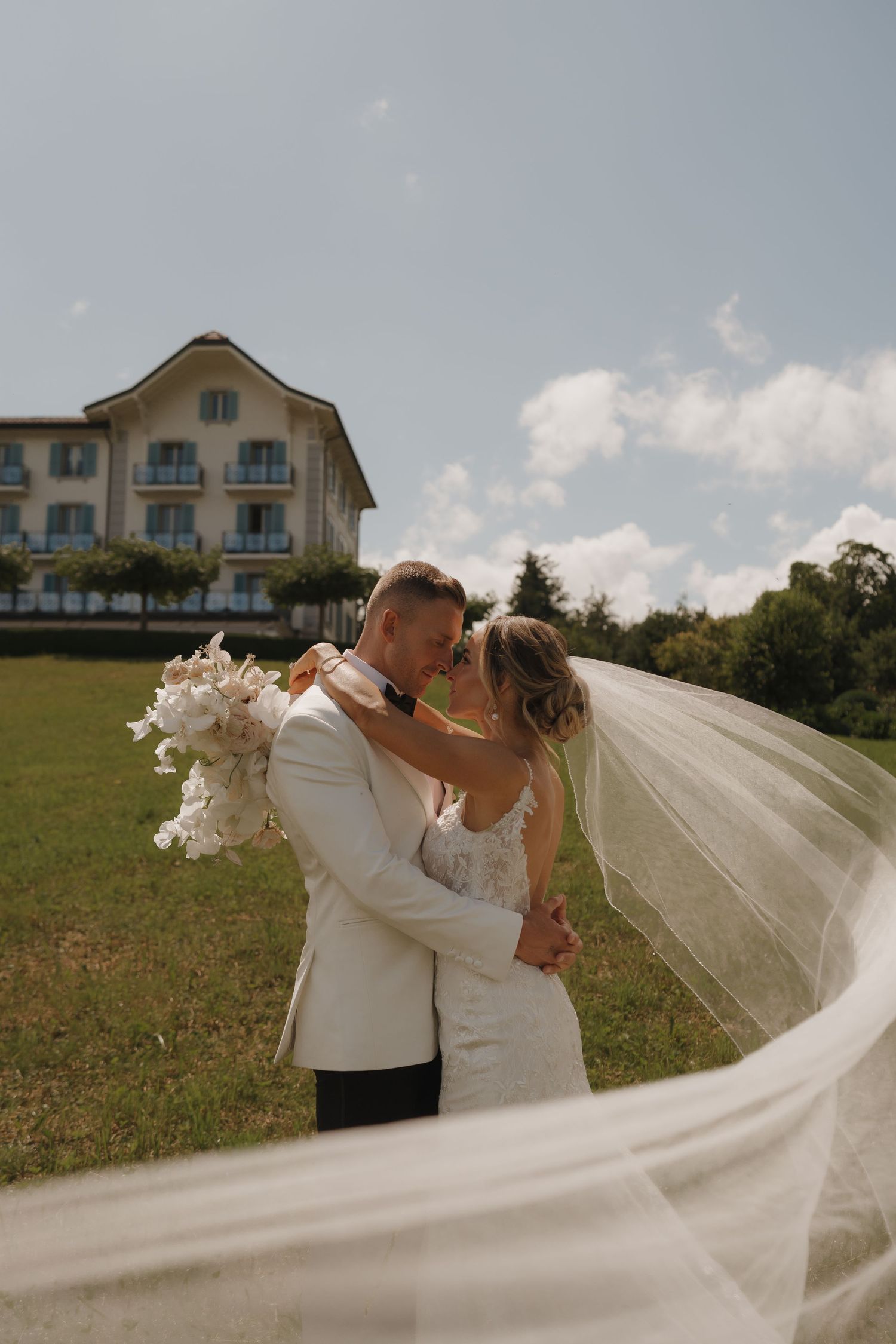 Wedding couple nearly kissing as the bride's veil flows in the wind, with a elegant villa building behind them.