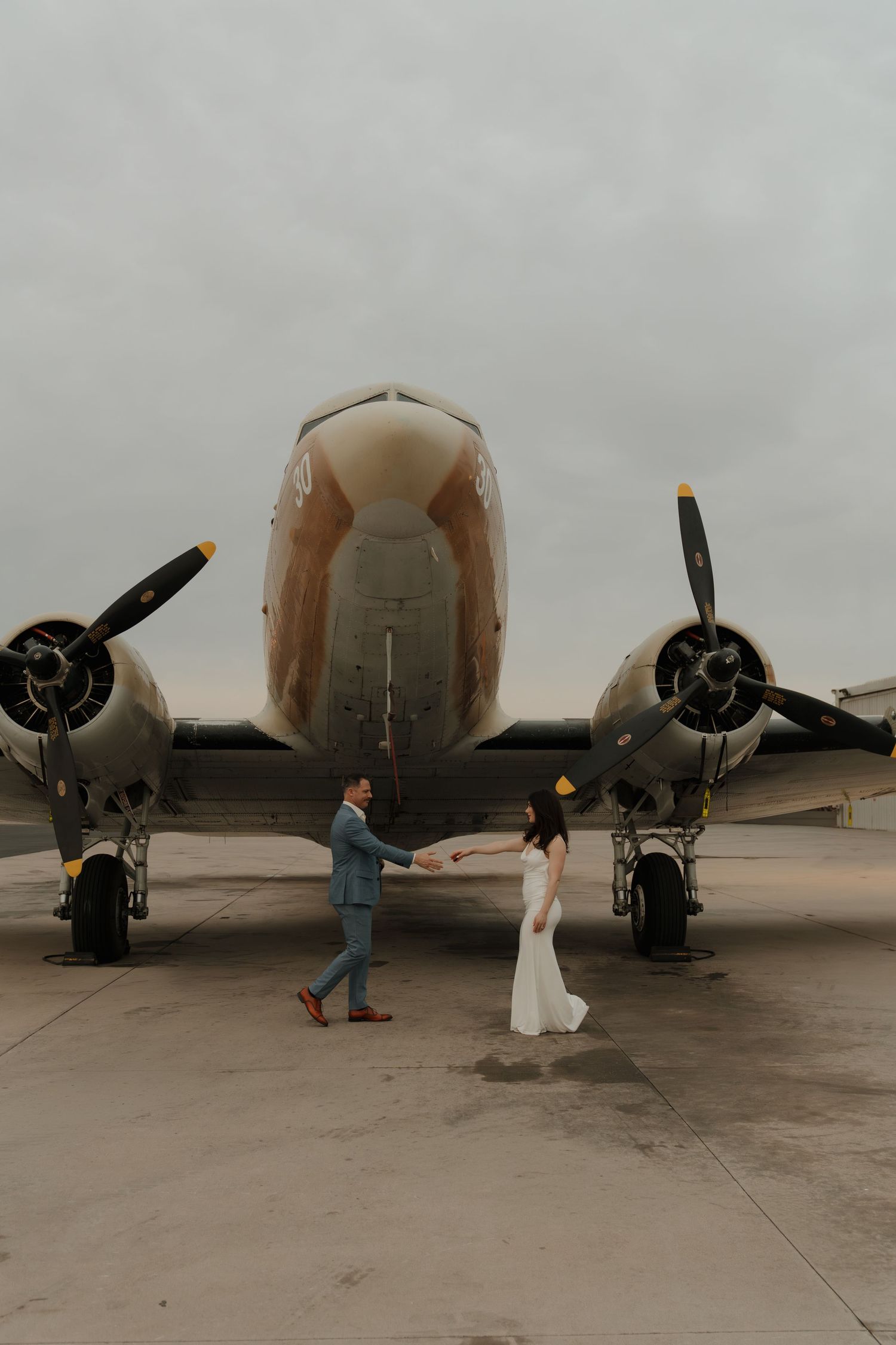 Bride and groom hold hands in front of a vintage propeller airplane on a tarmac during their wedding photo session.