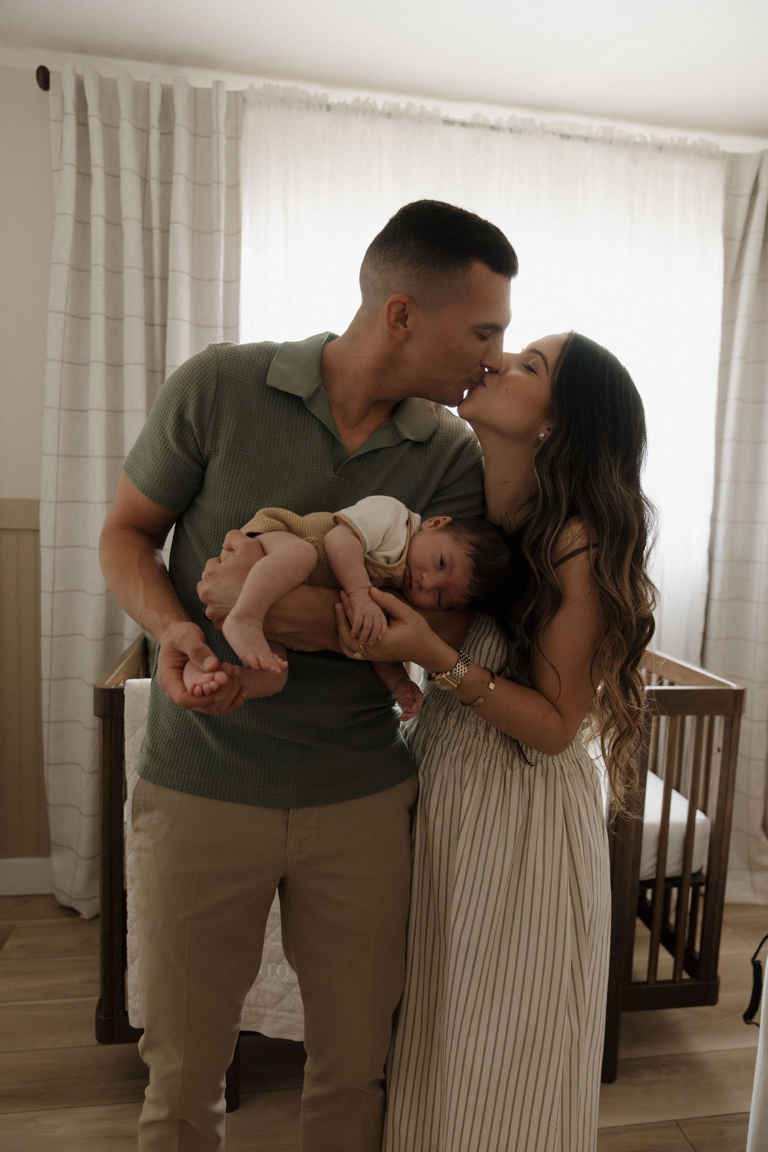 Couple kissing while holding sleeping newborn baby in front of wooden crib.