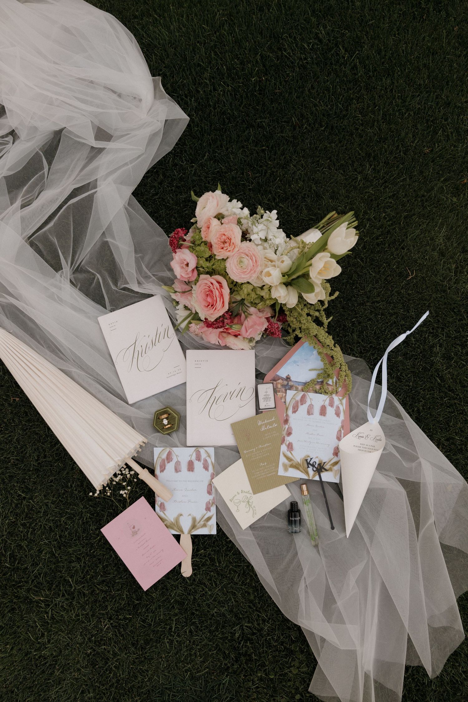 Elegant wedding flat lay with pink floral bouquet, invitation suite, veil, and accessories on green grass.