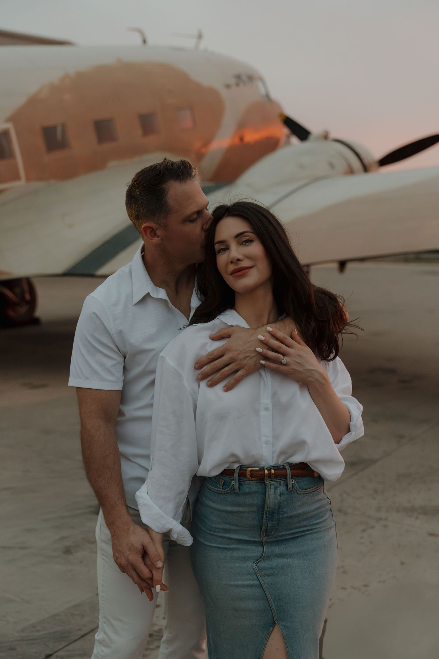 Romantic couple embracing in front of a vintage airplane at sunset, man kissing woman's cheek outdoors.