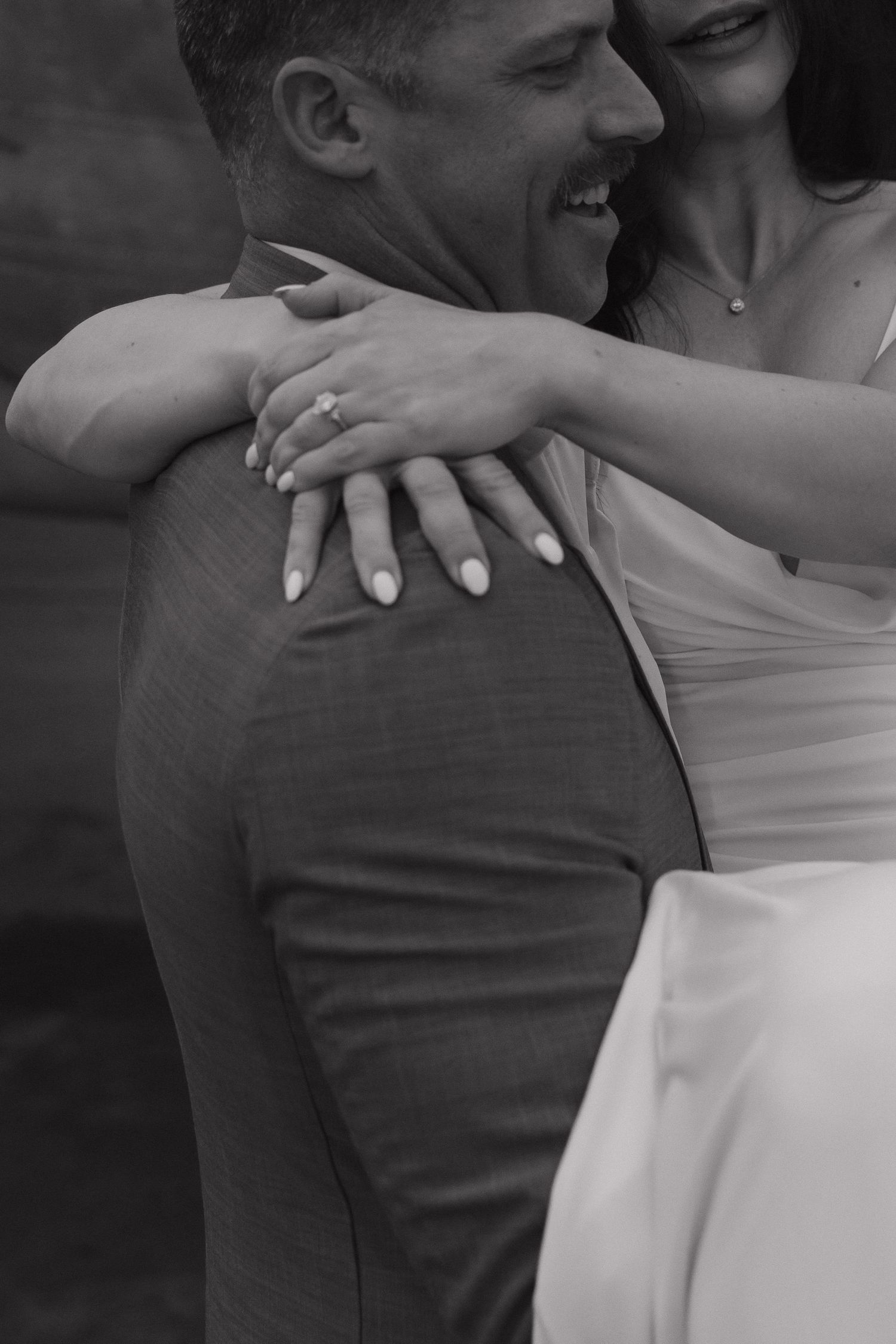 Black and white wedding photo of groom laughing as bride embraces him from behind, showing diamond engagement ring.