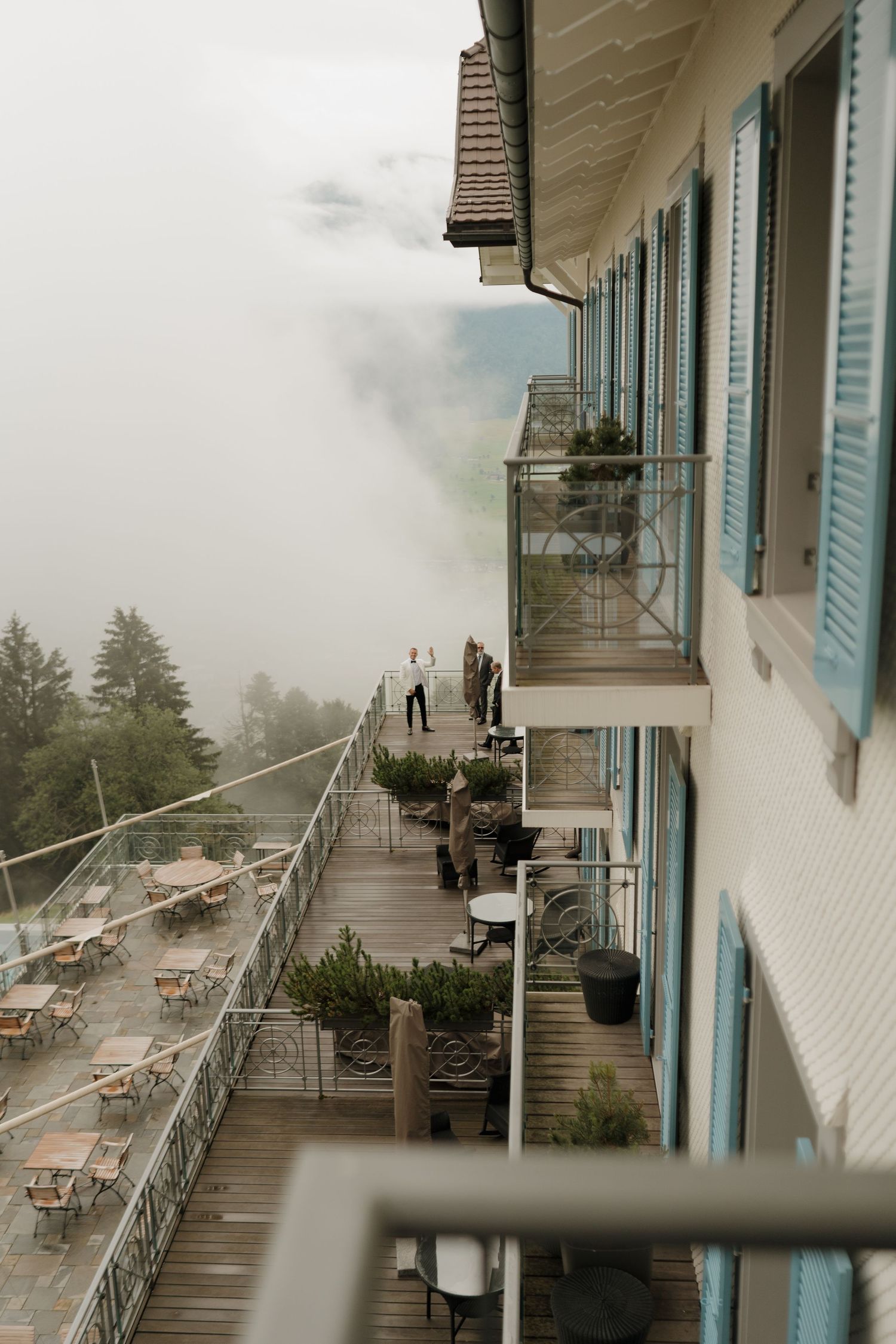 Misty mountain hotel terrace with blue shutters, outdoor dining area, and fog-covered alpine landscape.
