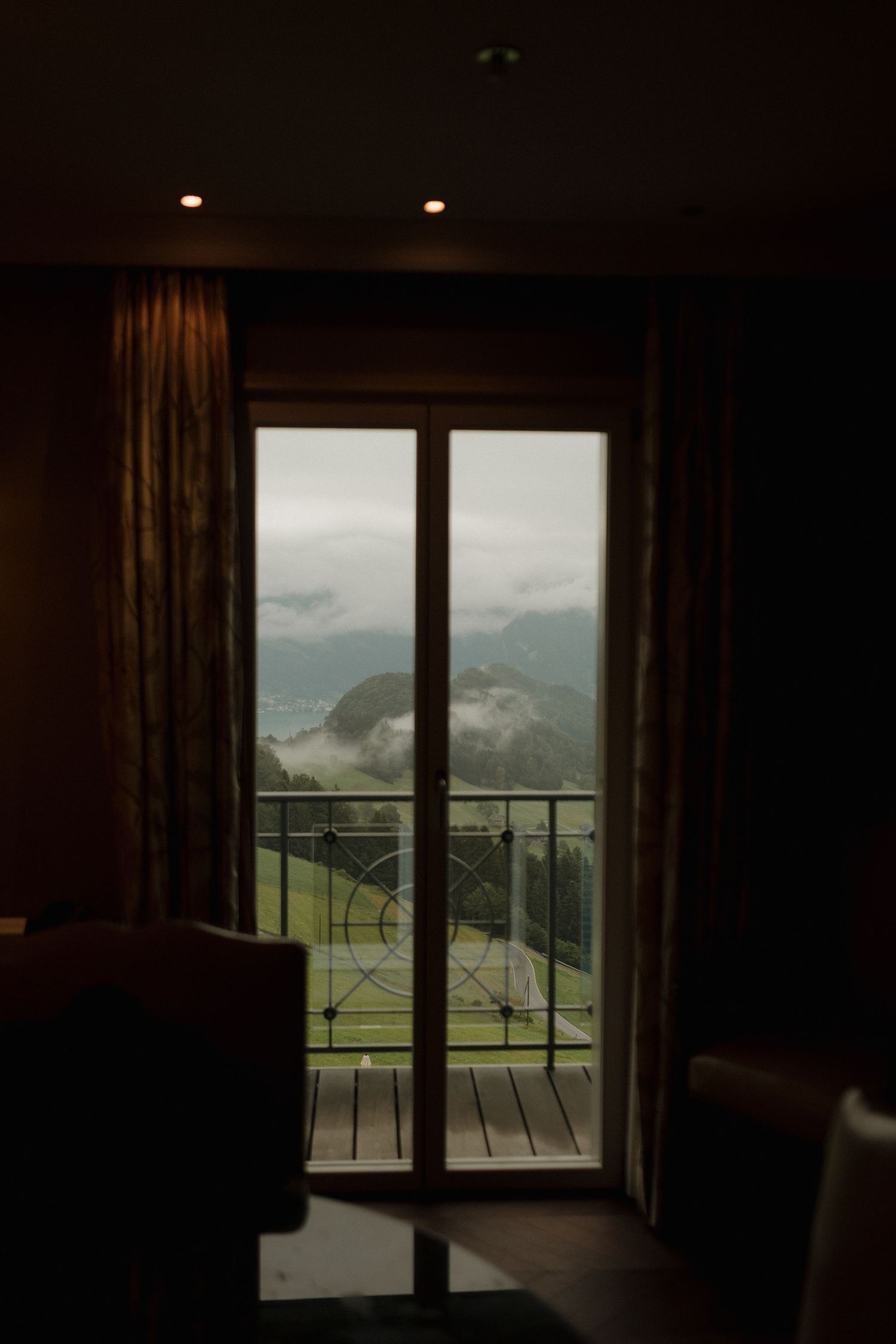 Dark hotel room with glass balcony door revealing misty mountain landscape view outside.
