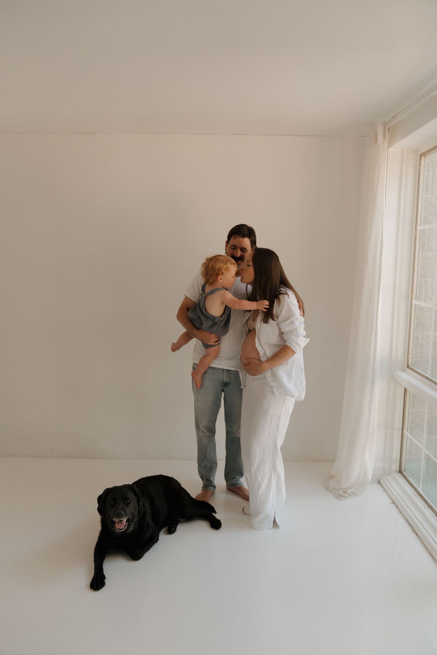 Family portrait of couple kissing baby in bright white studio with black Labrador resting on floor nearby.
