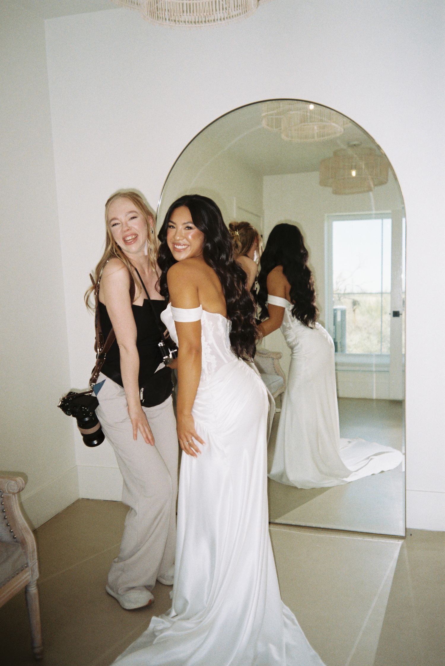 Bride in white off-shoulder gown poses with photographer near arched mirror in bright bridal suite.