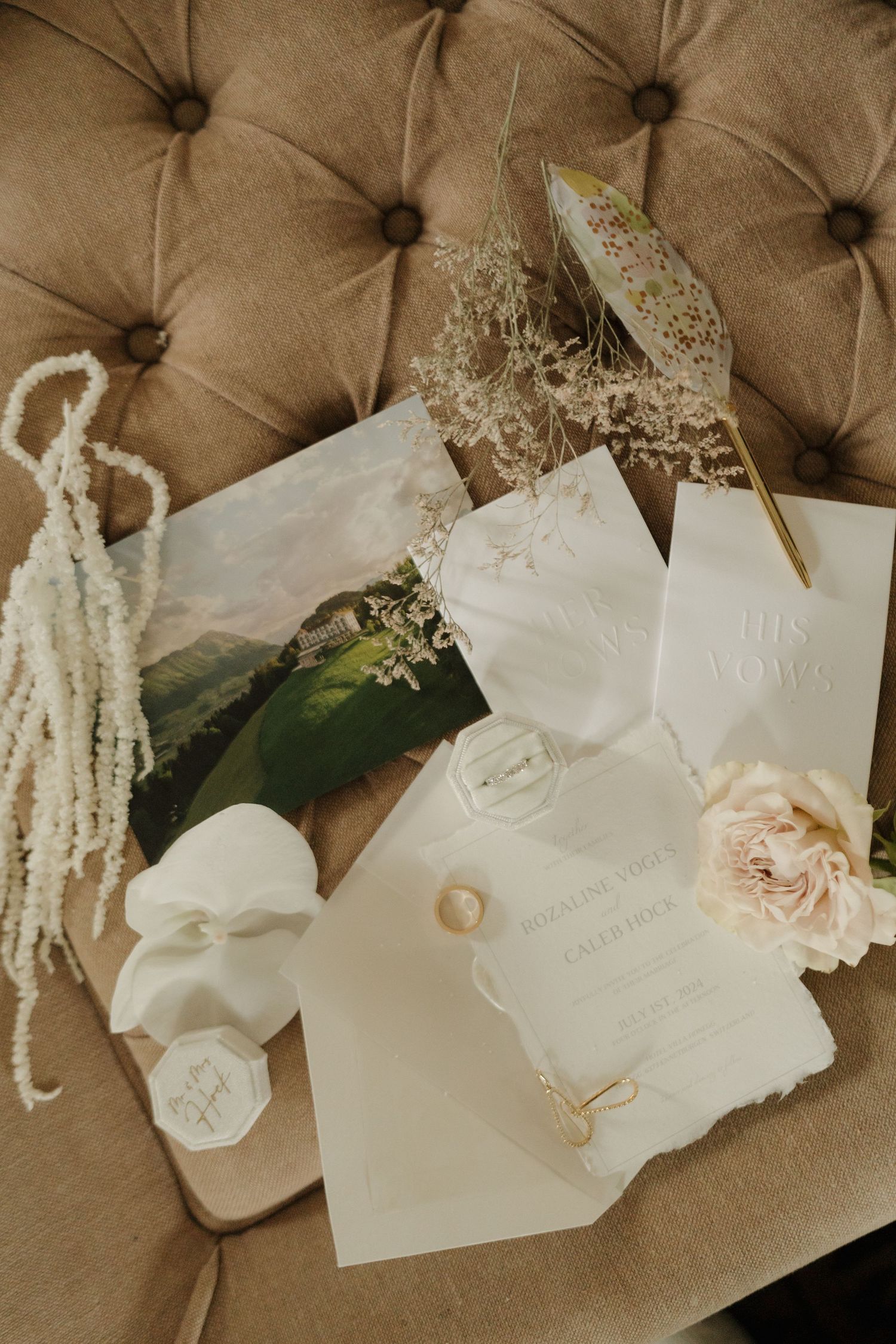 Elegant wedding stationery flat lay with rings, dried florals, and rose on tufted beige surface.