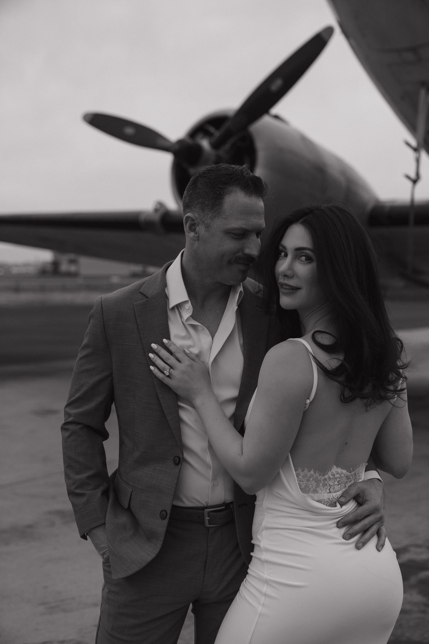 Elegant couple poses in black and white near vintage propeller plane, woman in white dress, man in suit.
