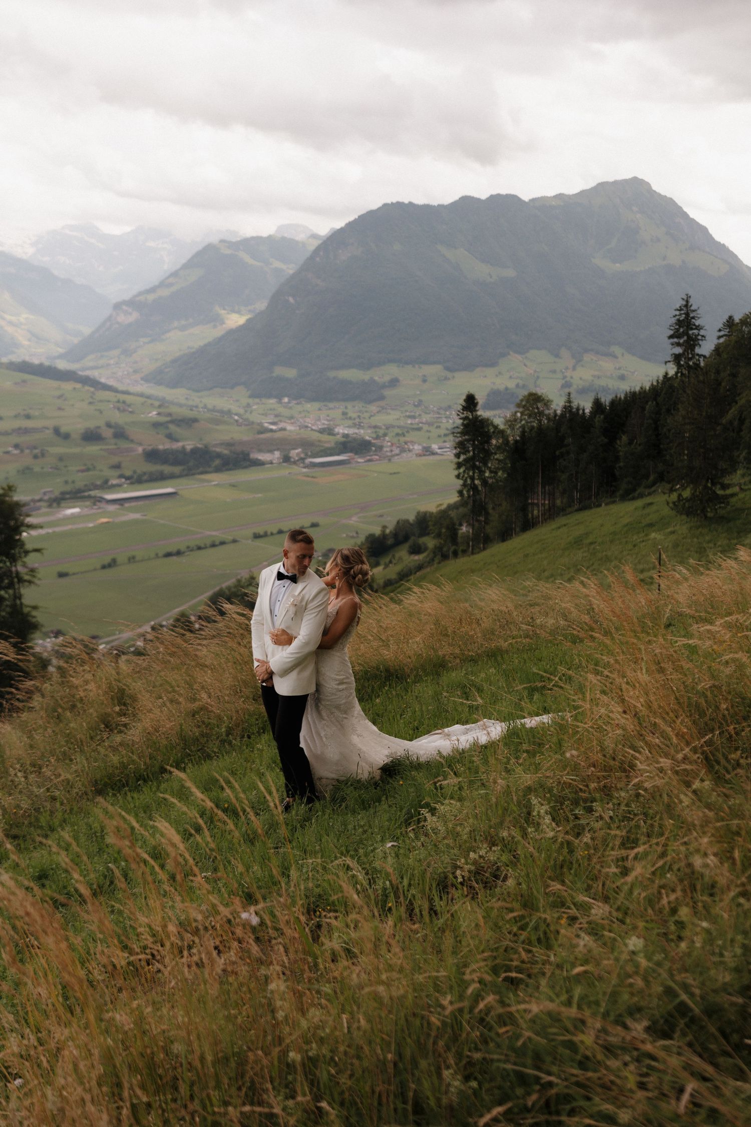 Bride and groom embracing on Alpine meadow with stunning mountain valley backdrop on wedding day.