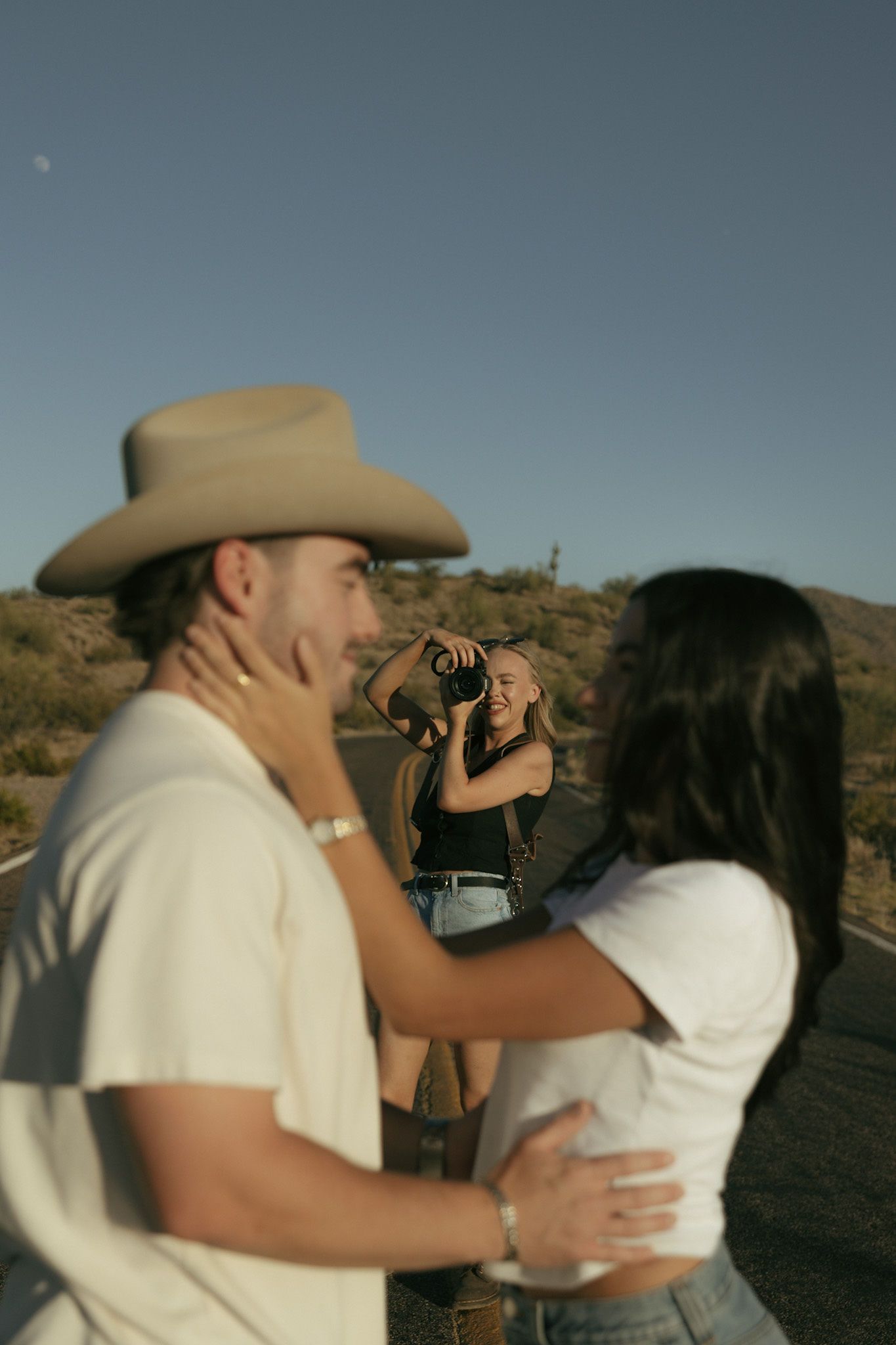 Photographer captures couple embracing on desert road, man in cowboy hat, woman in white top, arid landscape background.