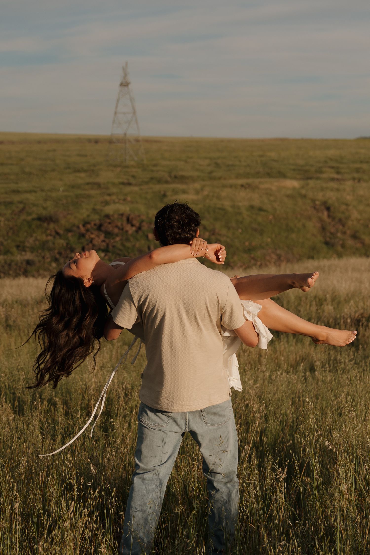 Man spinning woman in white dress in a green field at golden hour.