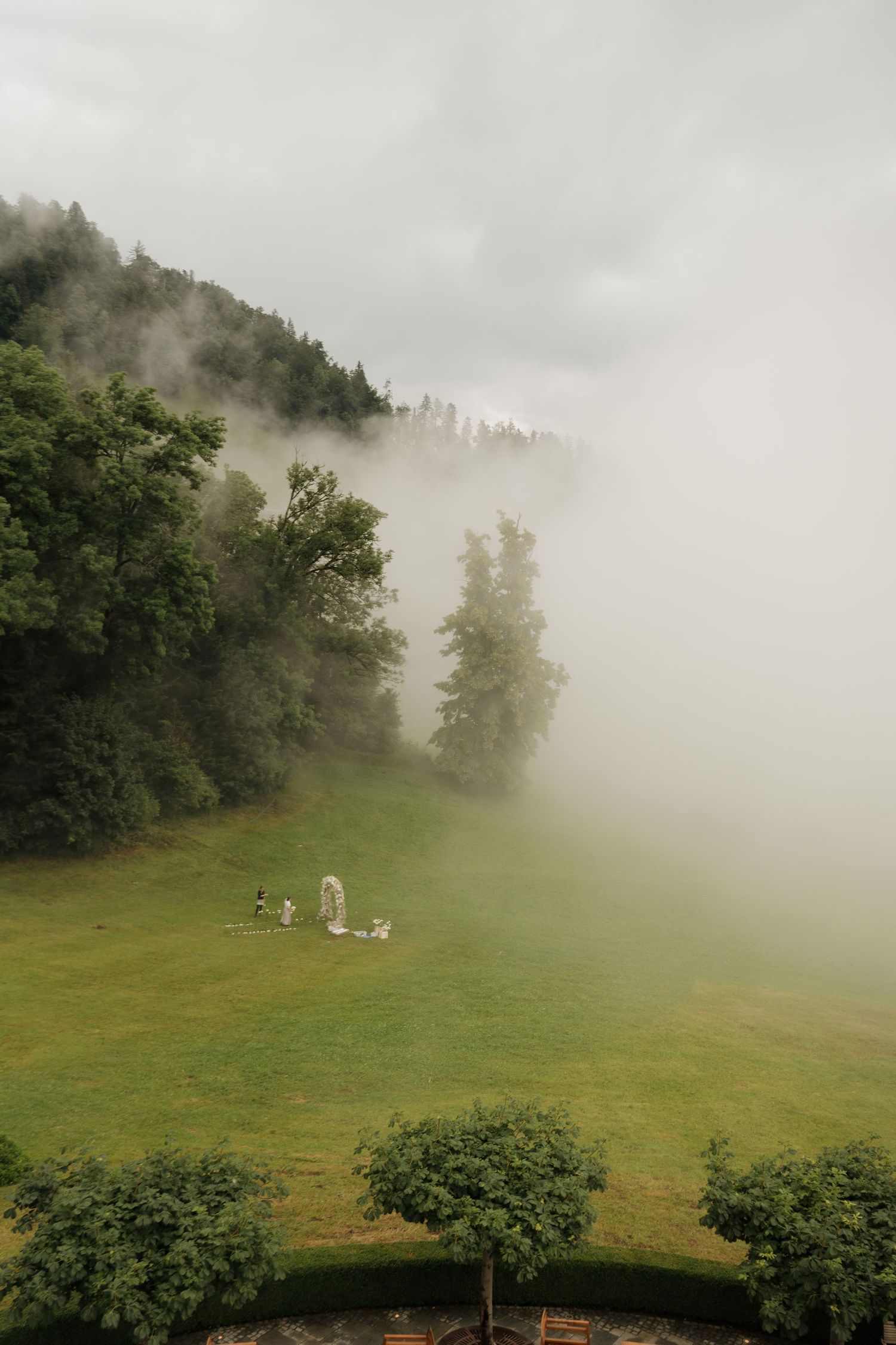 Foggy mountain landscape with tall evergreen trees and figures on lush green lawn below clouds.