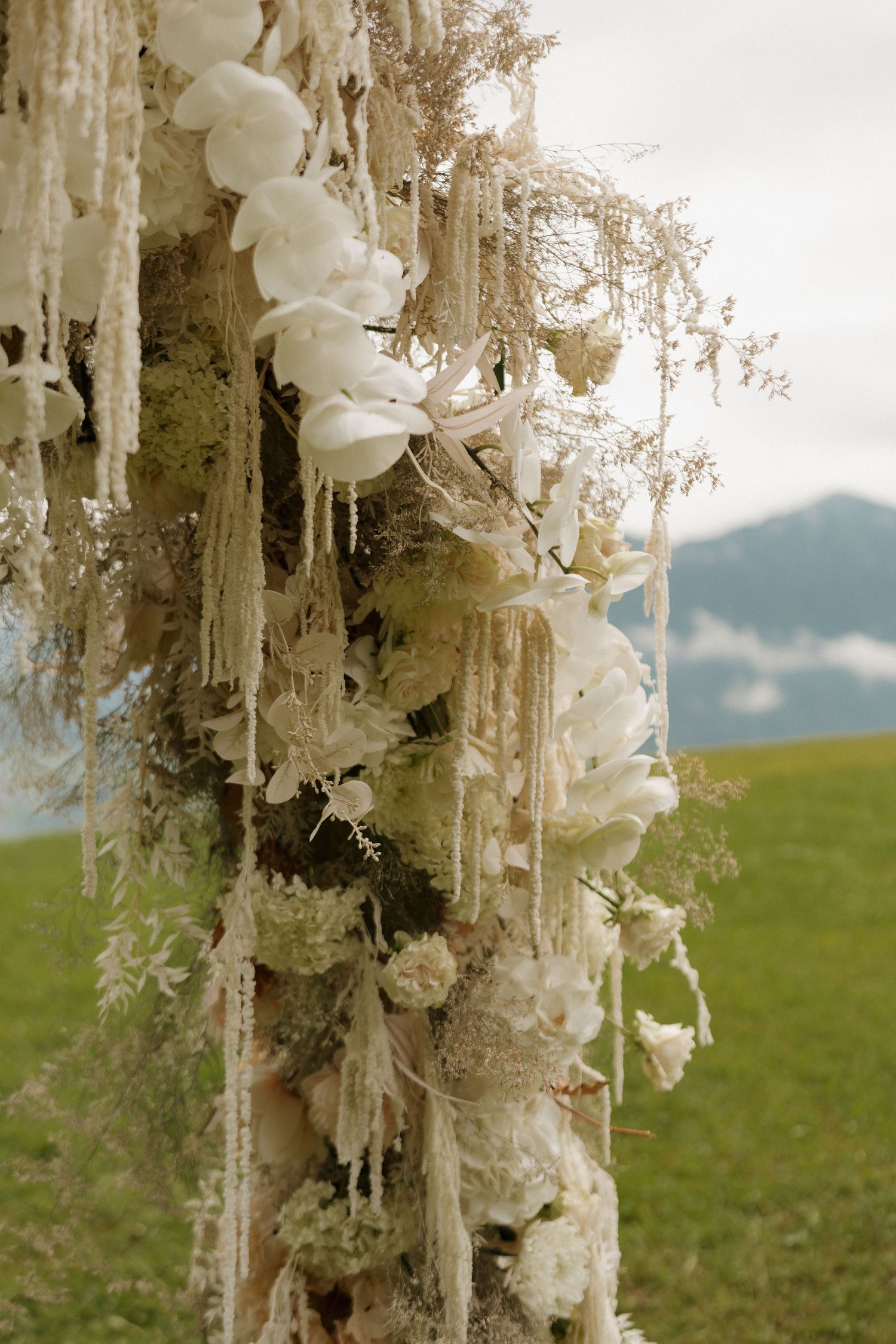 Elegant wedding arch column decorated with white orchids, roses, and hanging lace-like floral embellishments outdoors.