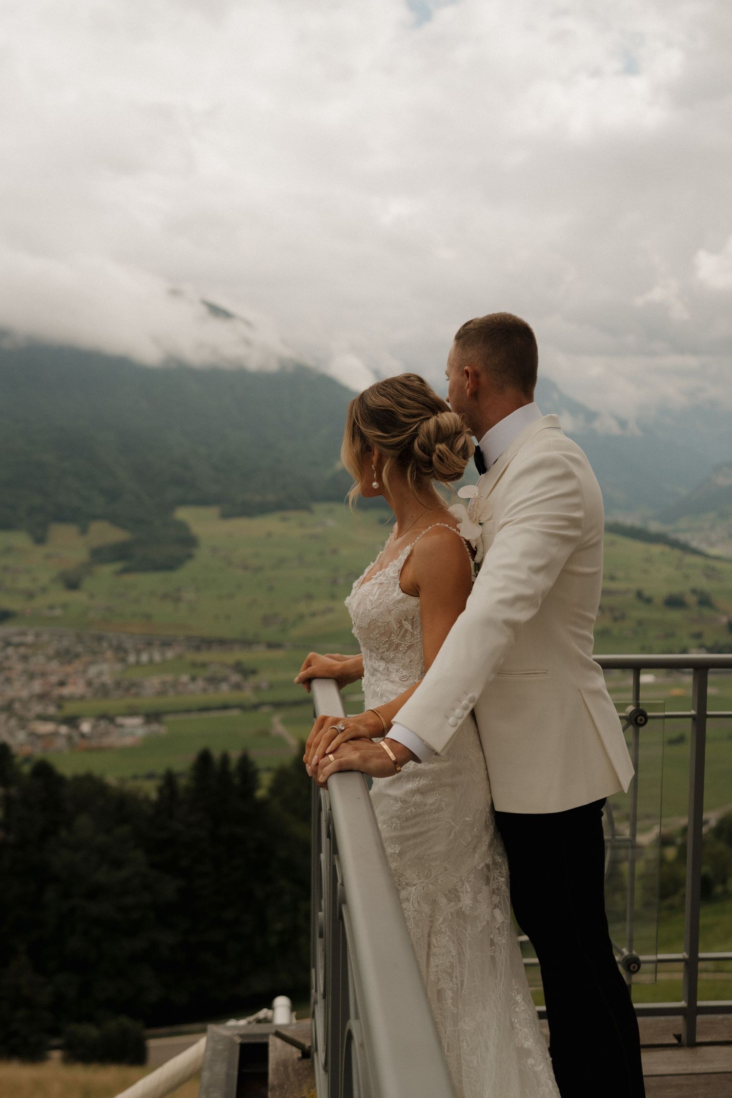 Bride and groom embracing on railing overlooking lush green alpine valley on their wedding day.