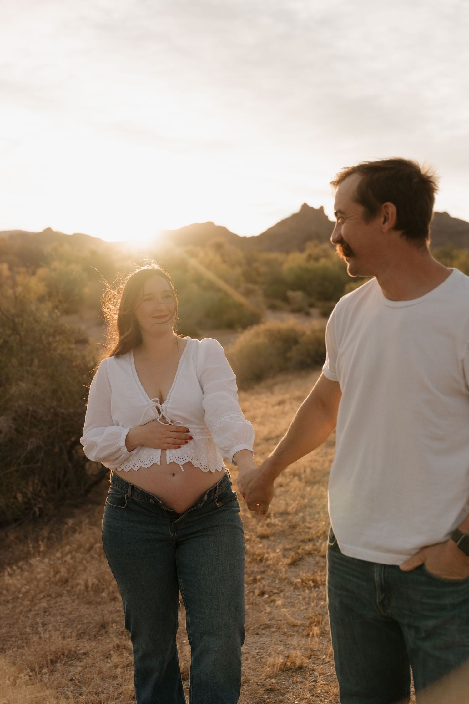 Pregnant woman in white top cradles baby bump while holding hands with partner at golden hour sunset.