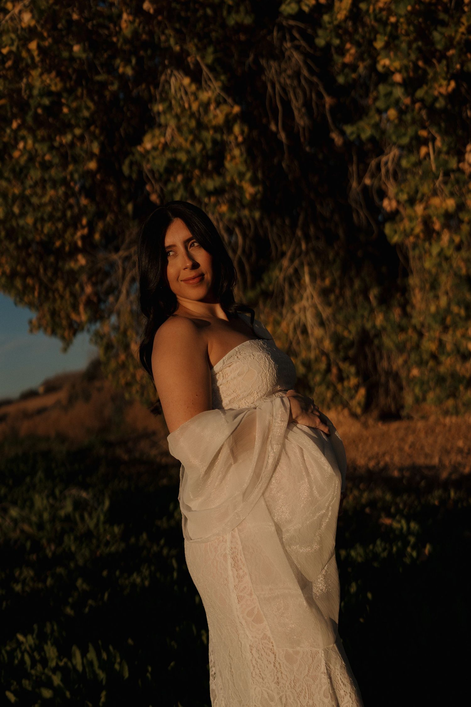 Pregnant woman in white lace dress holding baby bump under golden trees at sunset.
