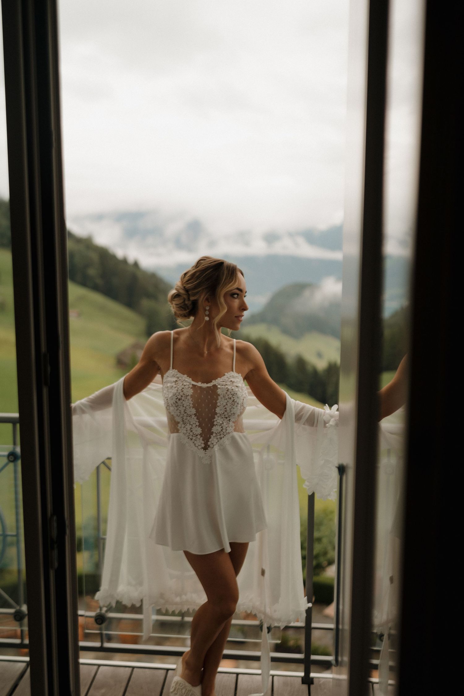 Bride in white lace lingerie and robe standing on a balcony doorway with scenic mountain views behind her.