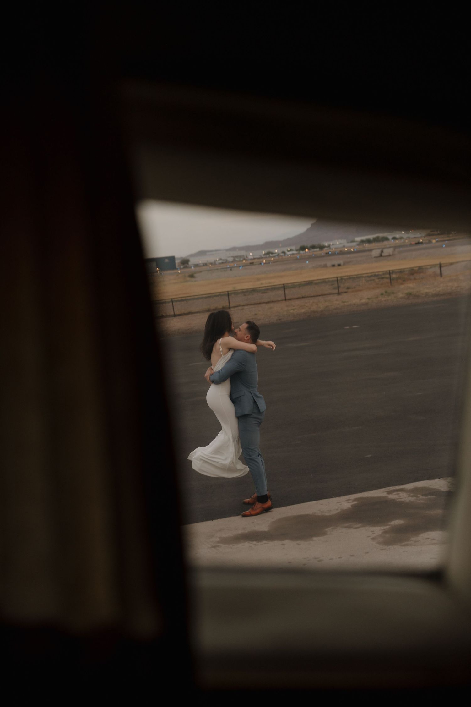 Bride and groom embracing outdoors near a runway, viewed through a dark window frame.