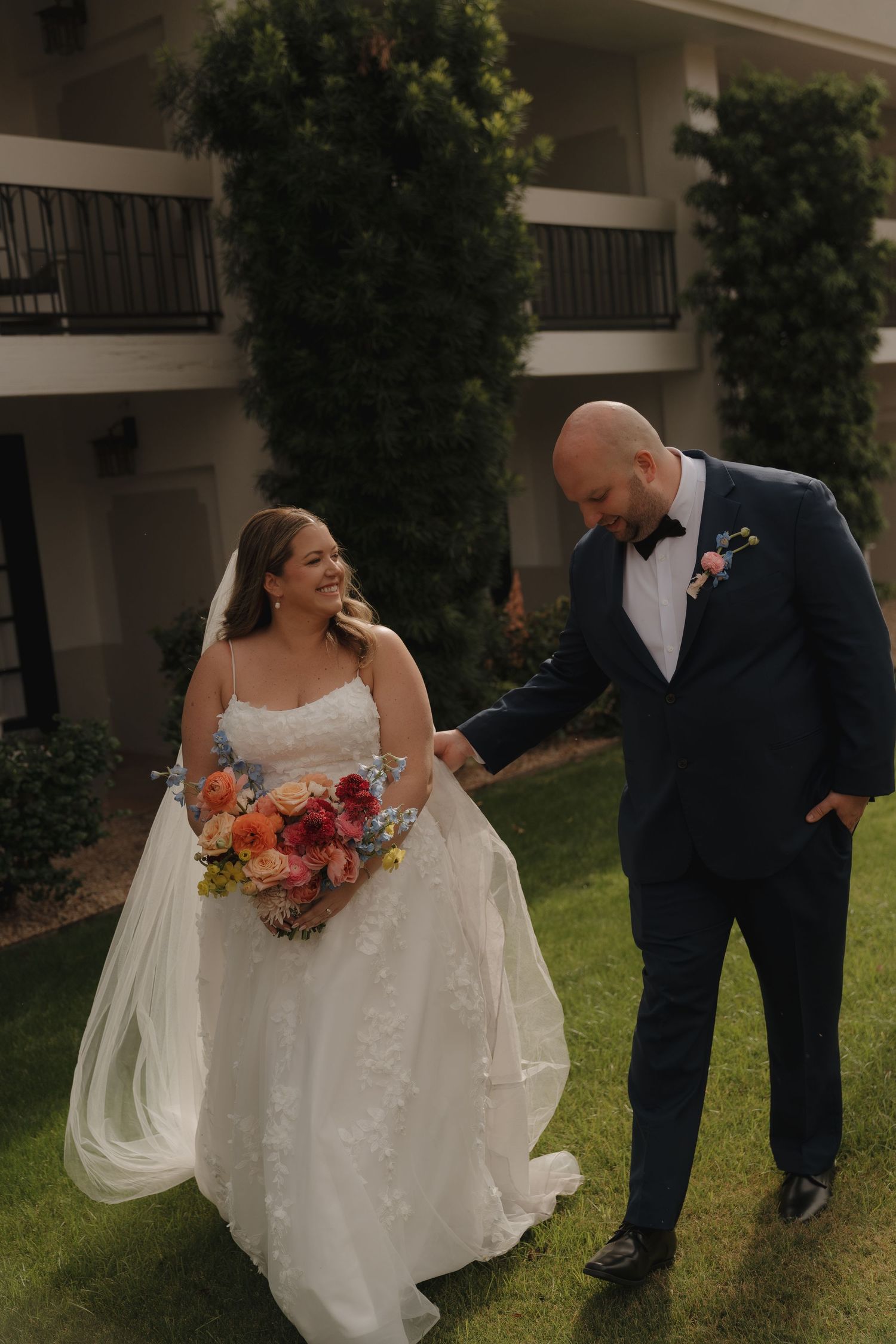Smiling bride in lace gown holding colorful bouquet walks with groom in navy tuxedo outdoors.