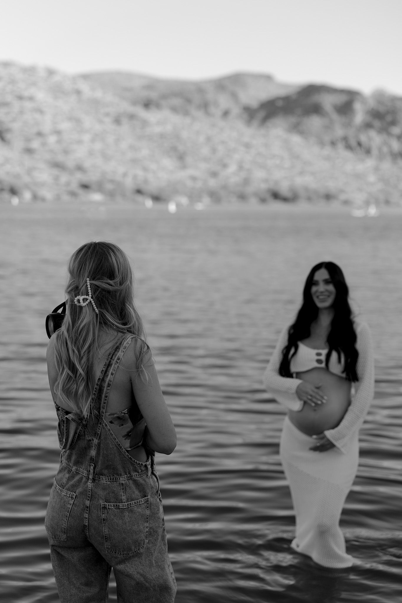 Black and white photo of two women standing in a lake, one pregnant in a white dress, with mountains in the background.