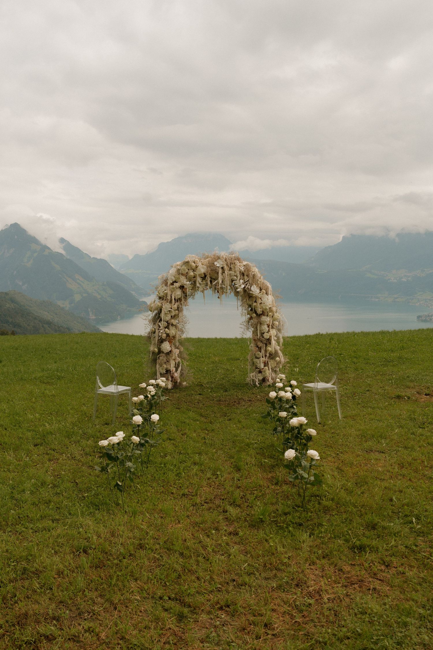 A stunning white floral wedding arch frames a breathtaking Alpine lake and mountain backdrop on a meadow.