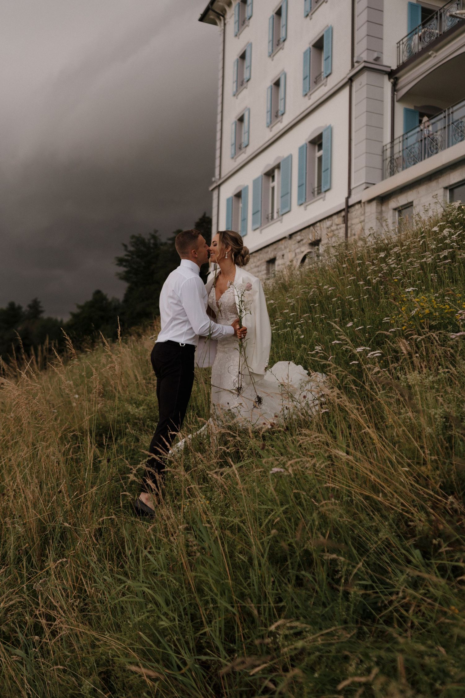 Bride and groom sharing a kiss in tall grass meadow with dramatic stormy sky behind them.