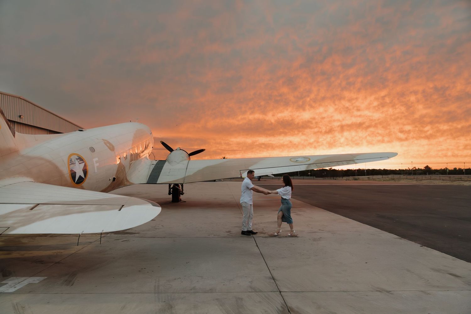 Two people stand beside a vintage white airplane on a tarmac during a stunning orange and red sunset.