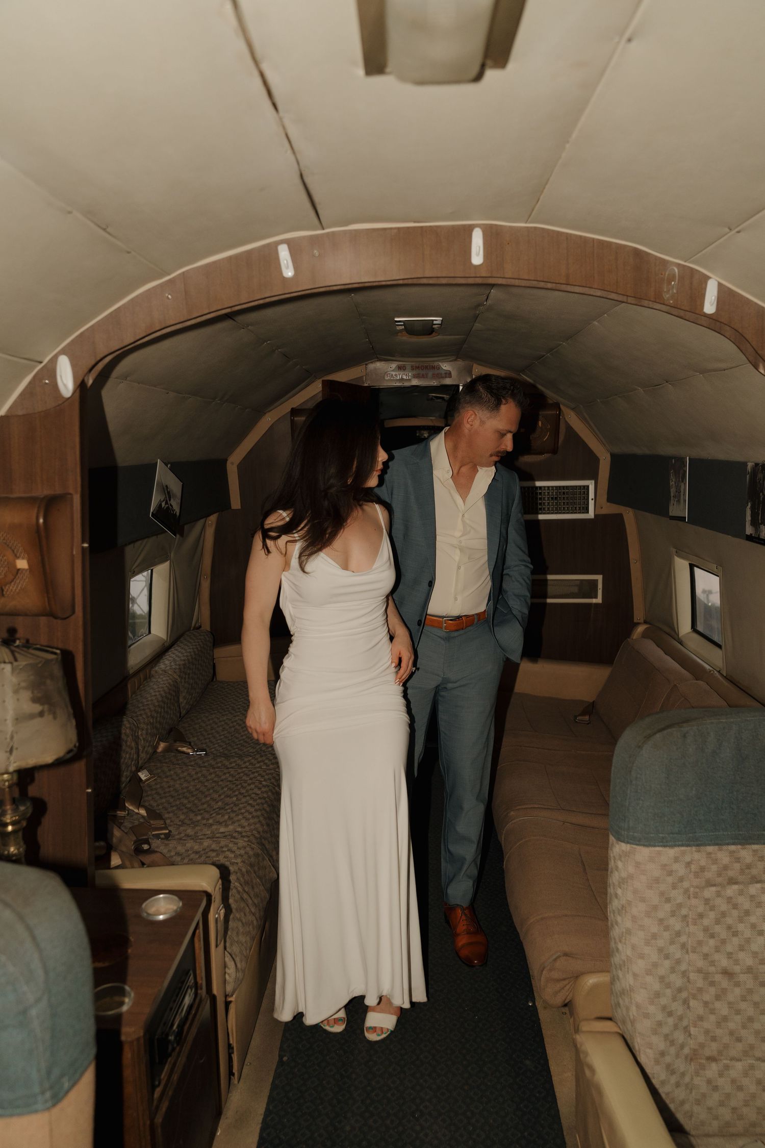 Couple in wedding attire walking through a vintage airplane interior, woman in white gown, man in blue suit.