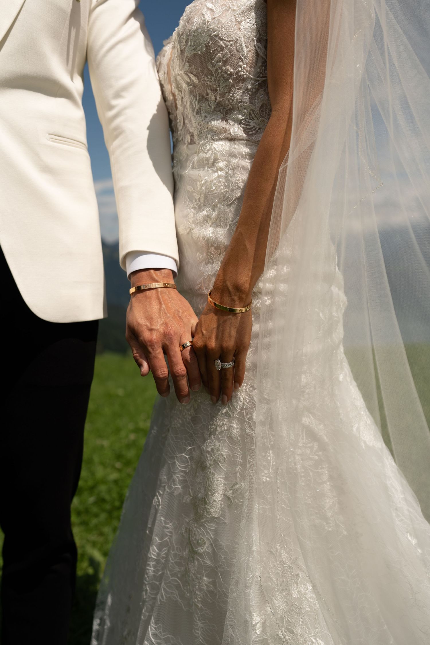 Close-up of newlyweds holding hands showing wedding rings and gold bracelets in elegant wedding attire.