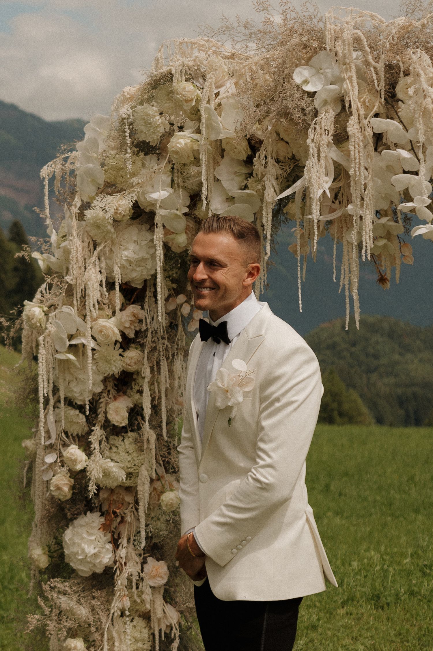 Smiling groom in a white tuxedo jacket stands beside an elaborate white floral wedding arch in a meadow.