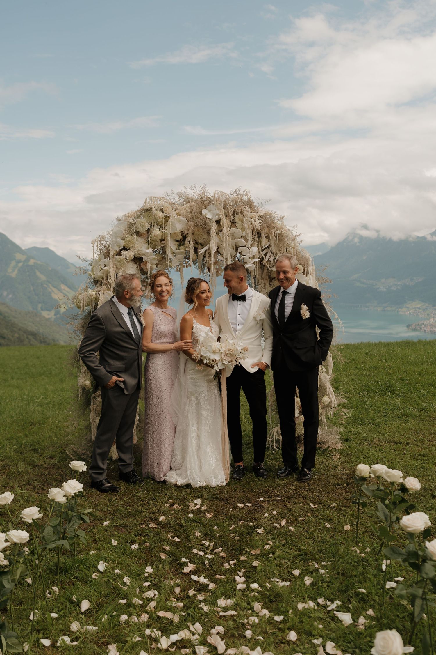 Wedding party posing under a floral arch in an Alpine meadow with lake and mountain views in the background.