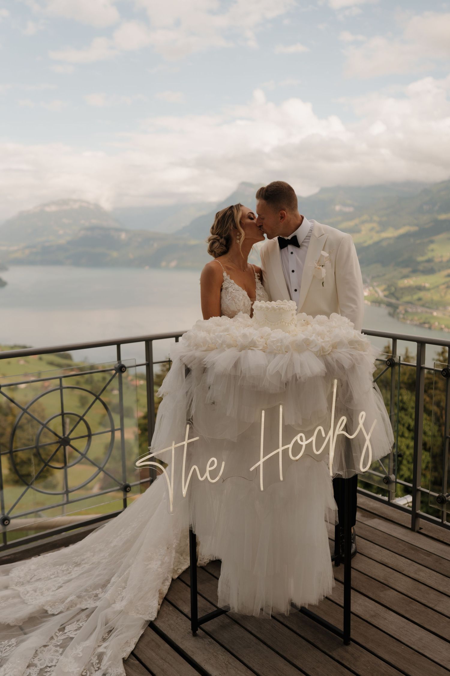 Bride and groom kissing on a scenic mountain balcony with lake views, beside a white floral wedding cake.