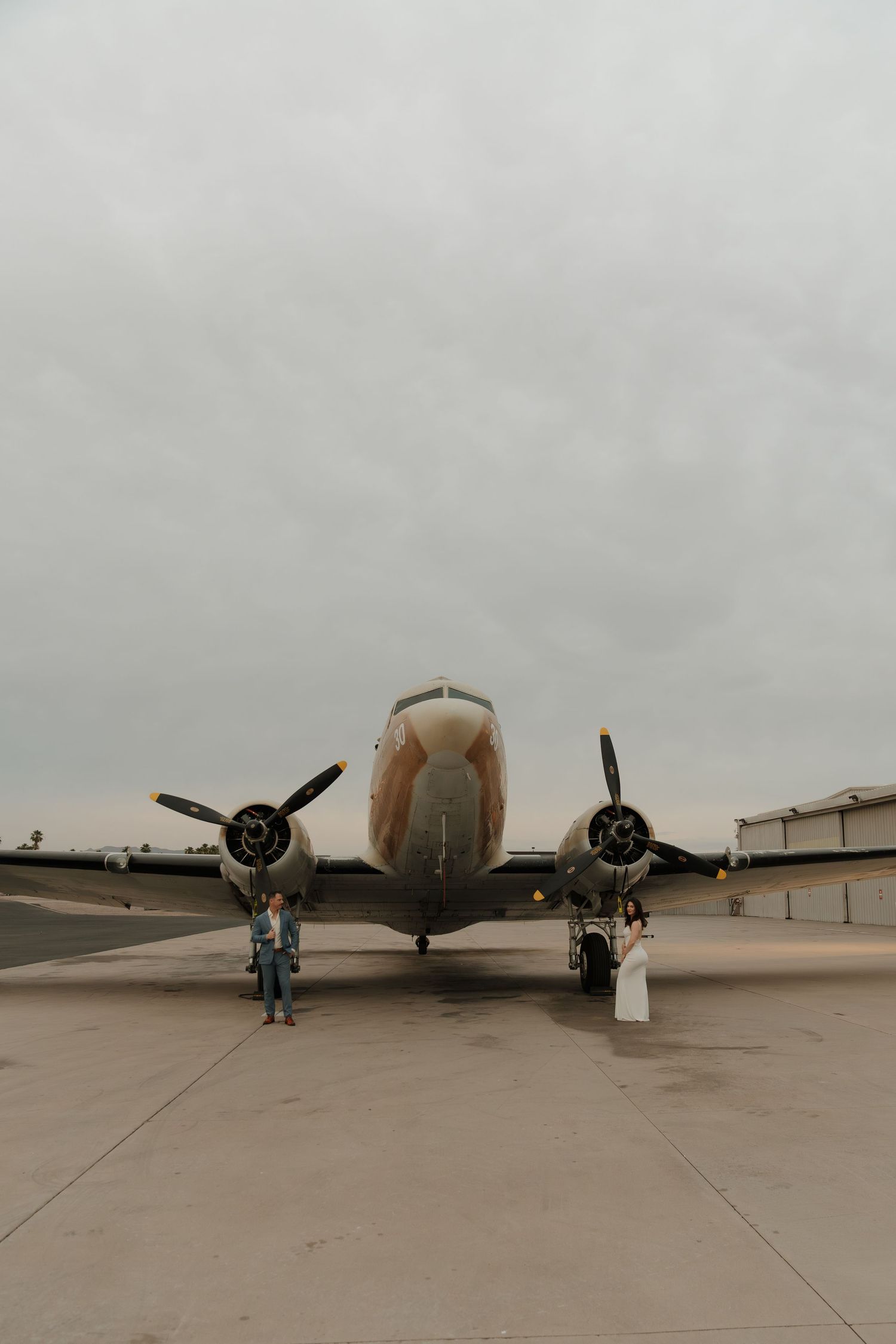 Couple poses before a vintage propeller aircraft on a tarmac under overcast skies in a wedding photo shoot.