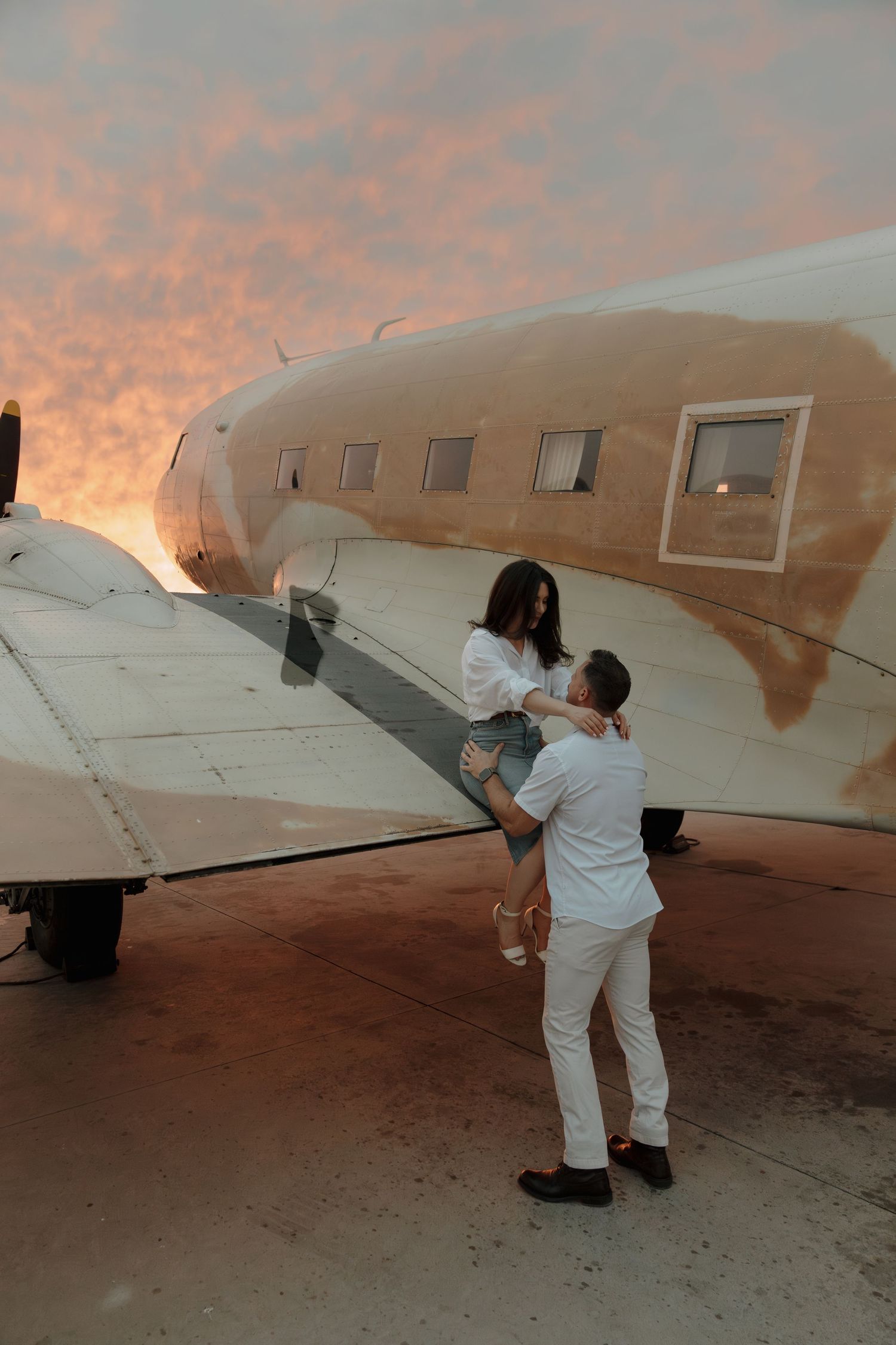Couple sharing romantic moment beside vintage airplane at sunset, man lifting woman near aircraft wing.