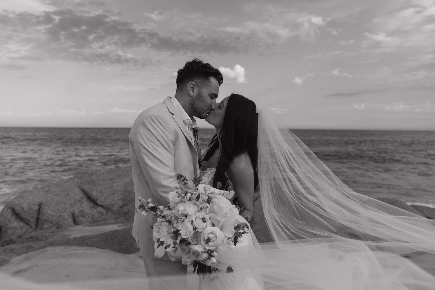 Black and white beach wedding photo of bride and groom kissing with ocean backdrop in Cabo.