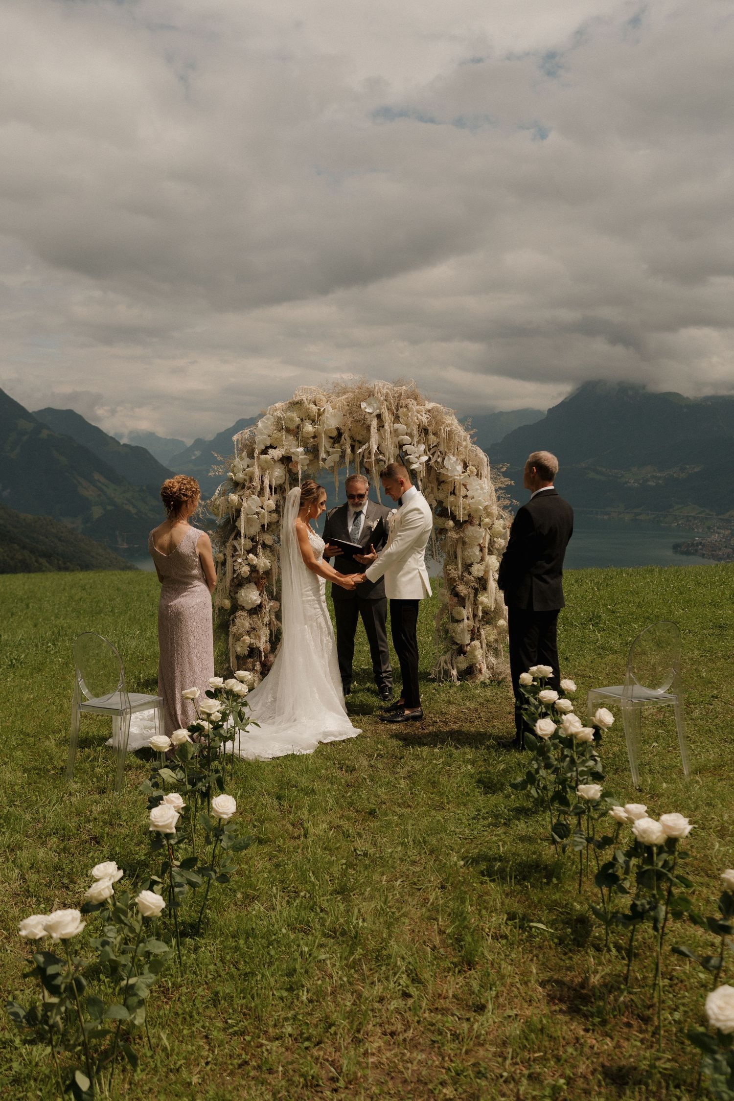 Bride and groom hold hands at the altar under a white floral arch with dramatic mountain scenery behind them.