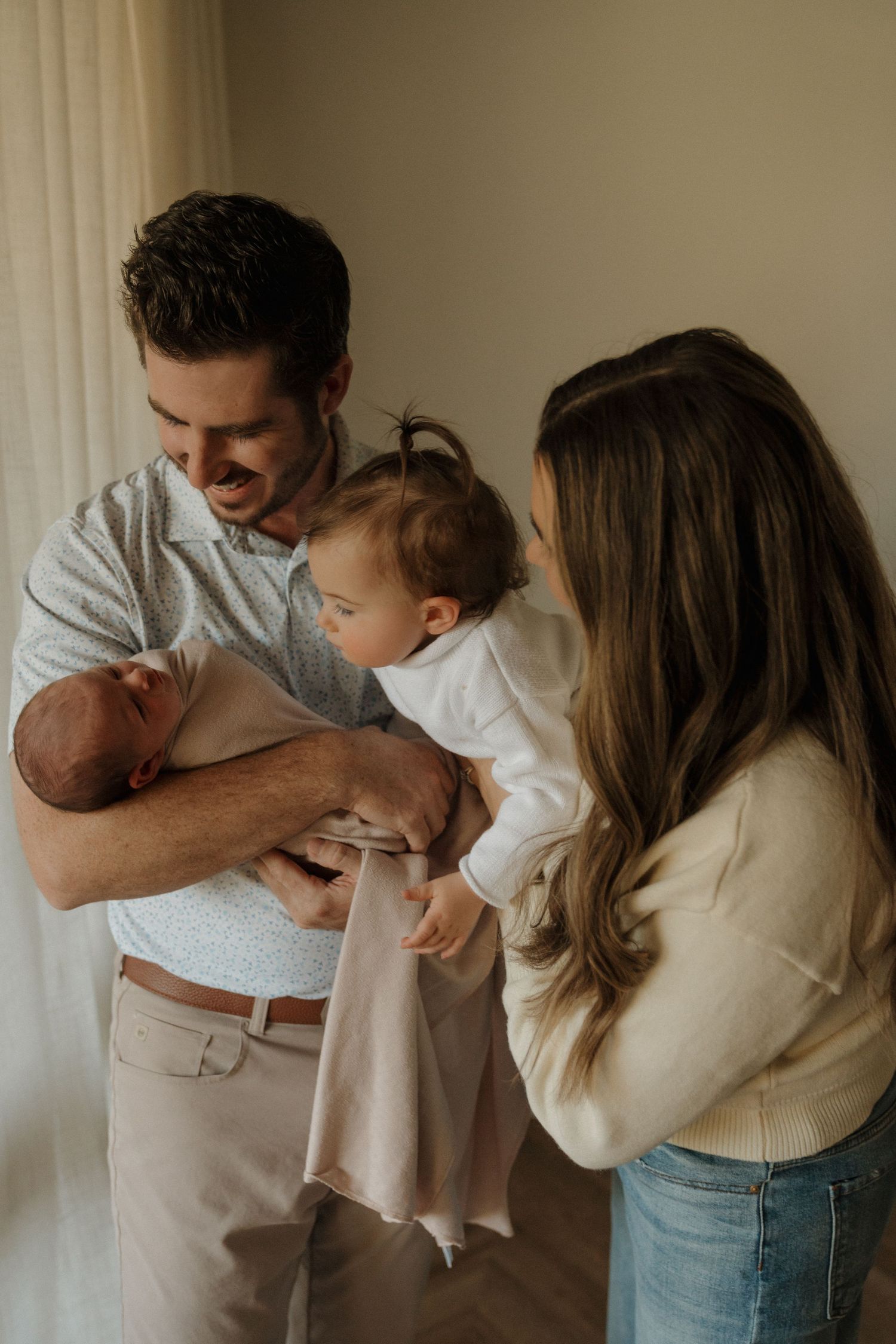 Father holding newborn while toddler reaches out, mother standing beside them indoors.