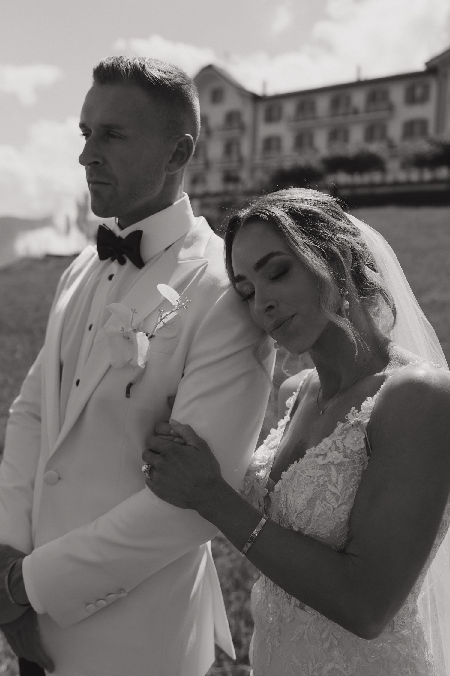 Black and white portrait of bride leaning on grooms shoulder in white tuxedo with historic building backdrop.