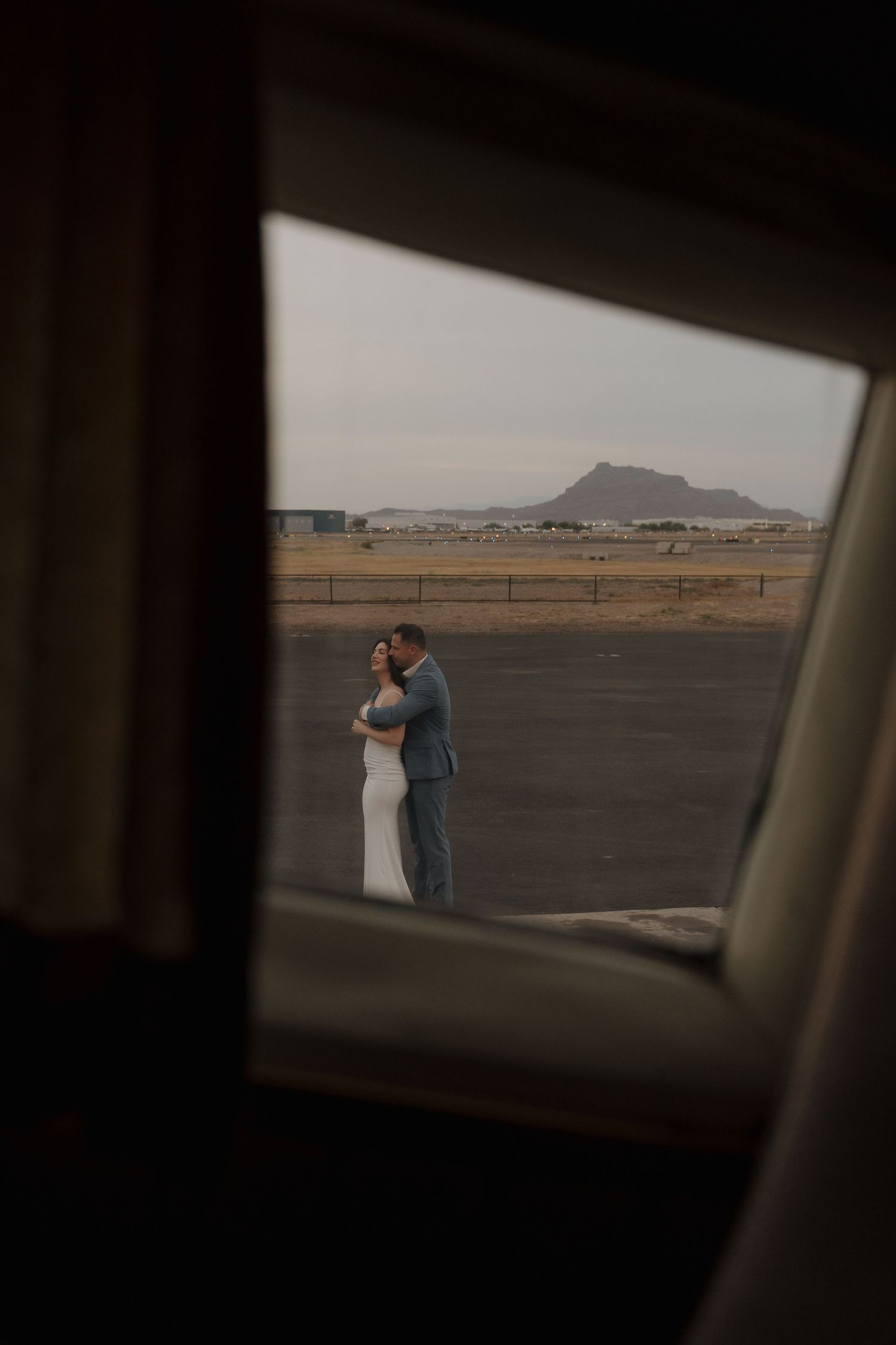 A couple in wedding attire embraces outdoors, framed by a window with a mountain landscape backdrop.