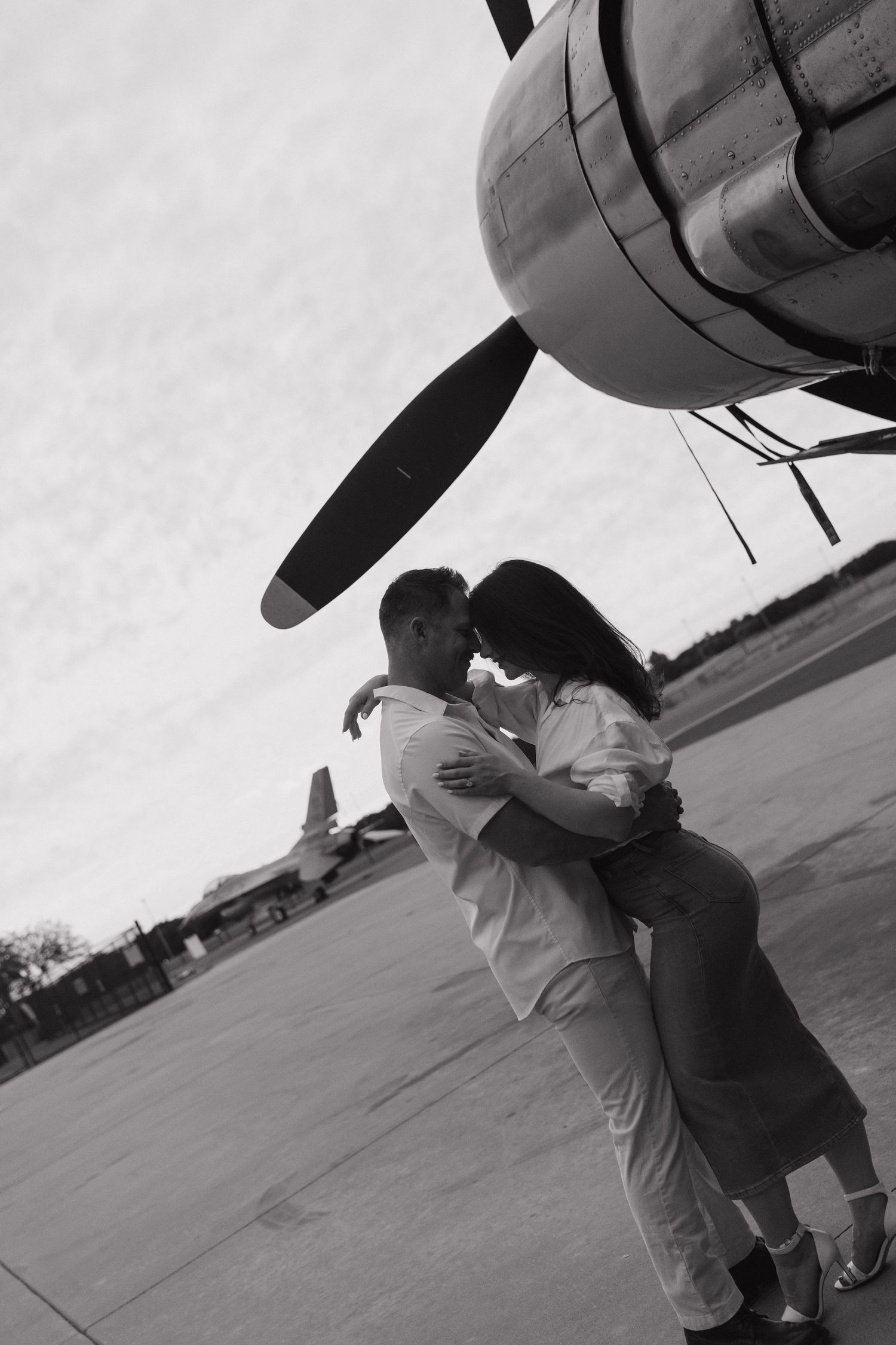 Black and white photo of a romantic couple embracing near a vintage airplane propeller on an airfield.