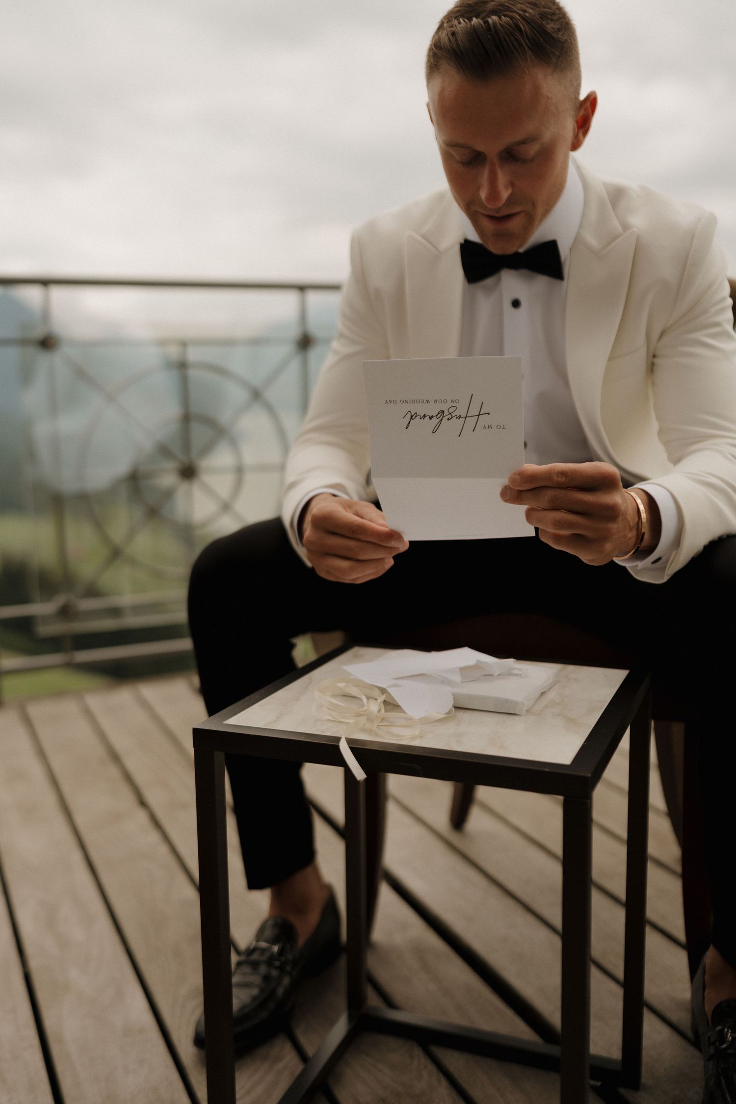 Groom in white tuxedo jacket reads wedding vow card on outdoor balcony with mountain views.
