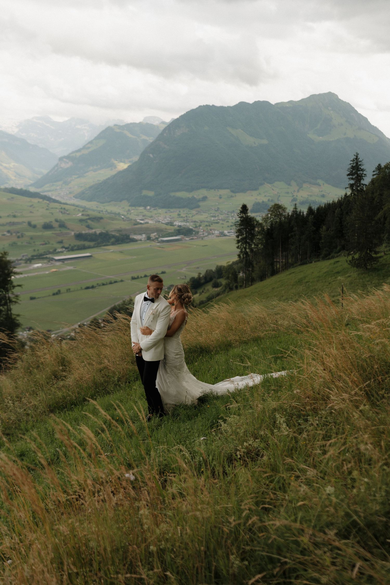 Wedding couple embracing on alpine meadow with stunning mountain valley backdrop on cloudy day.
