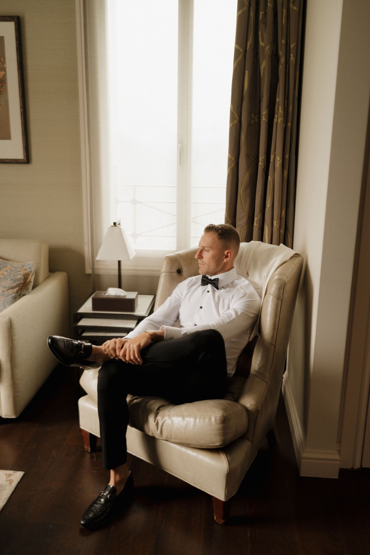 Dapper groom in white shirt and black bow tie relaxing in cream armchair by hotel window.