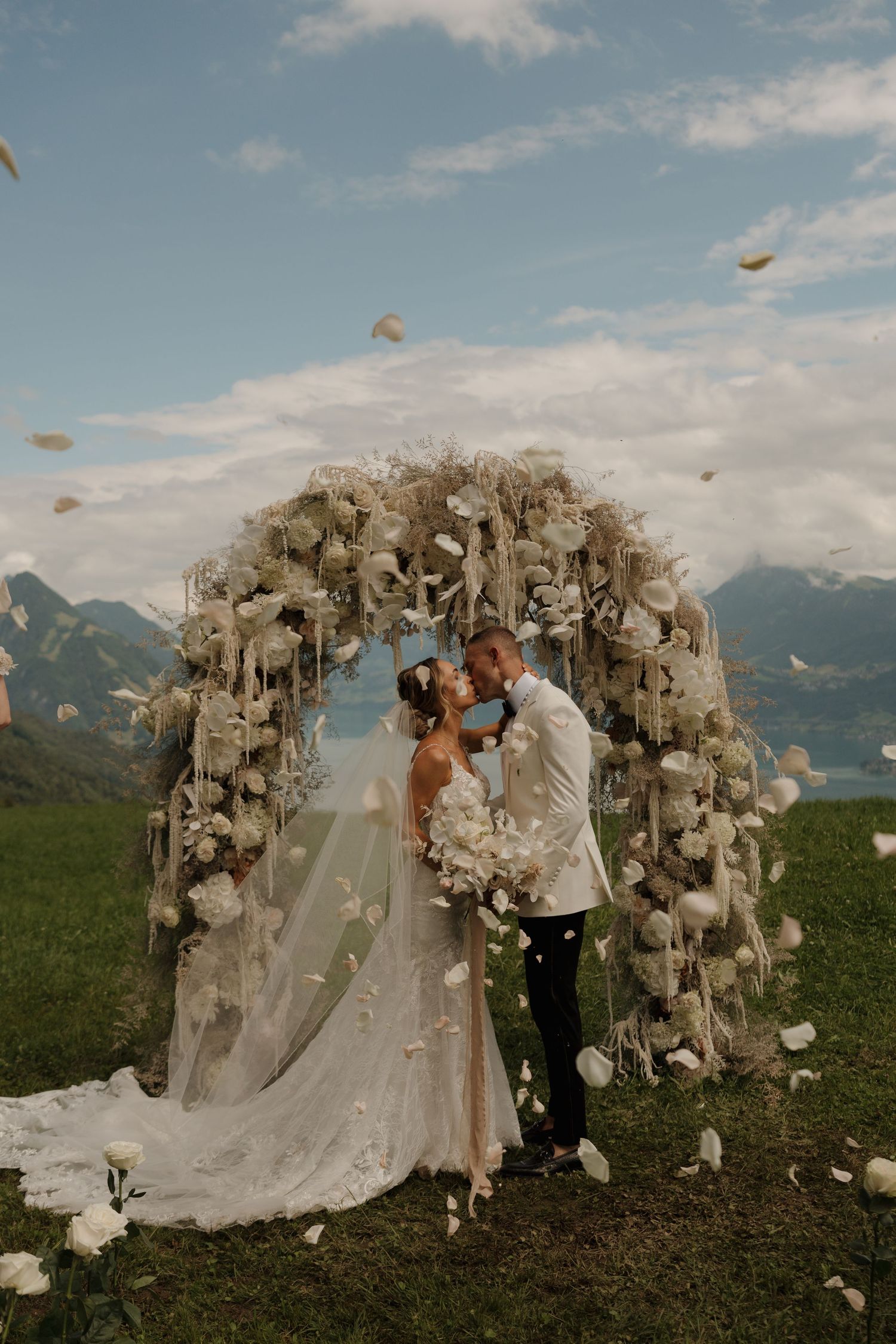 Bride and groom share a kiss under an elegant white floral arch with mountain lake views behind them.