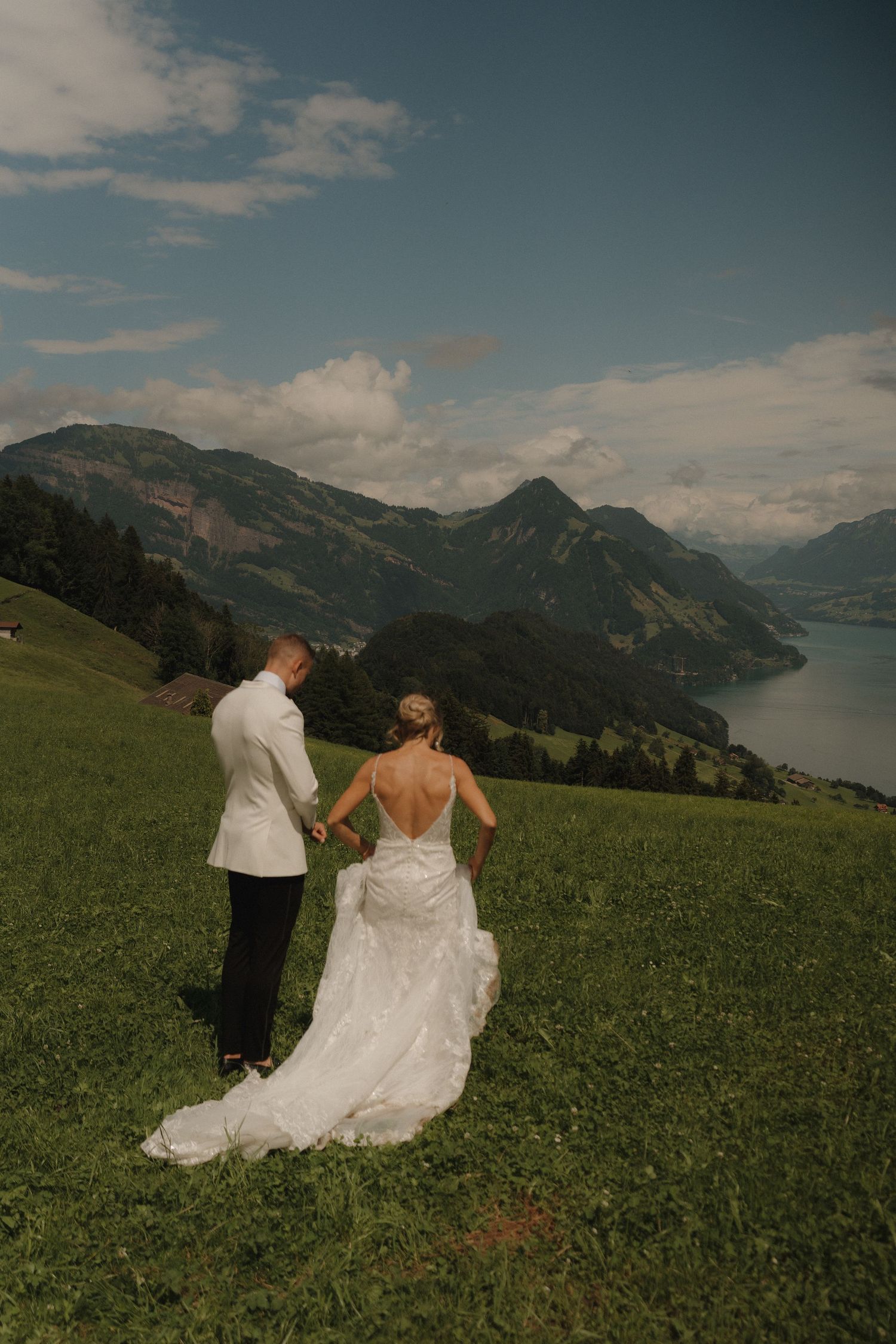 Bride and groom walk hand in hand toward stunning Swiss alpine lake and mountain panorama on sunny day.