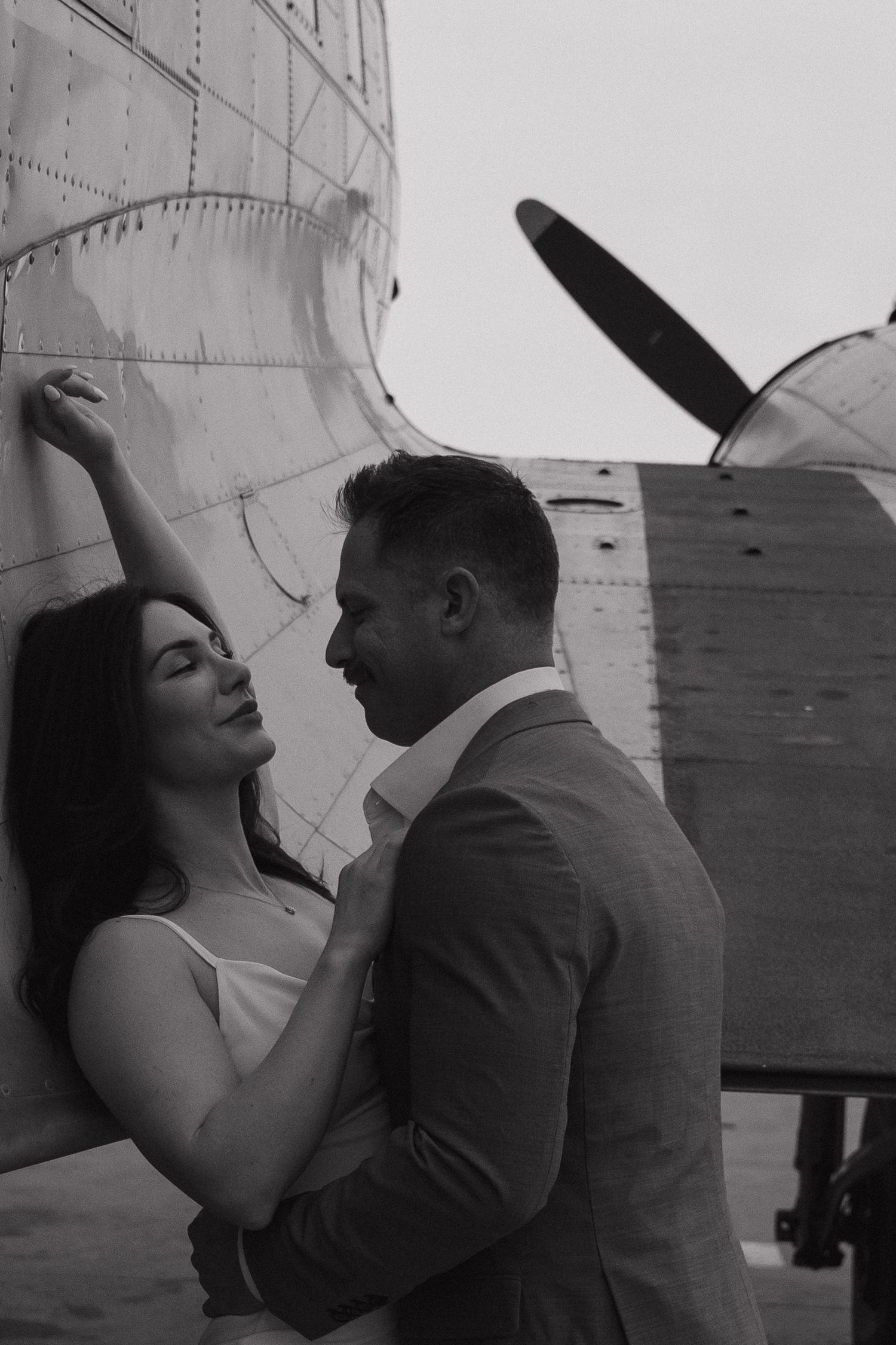 Black and white photo of a romantic couple embracing near a vintage propeller airplane.