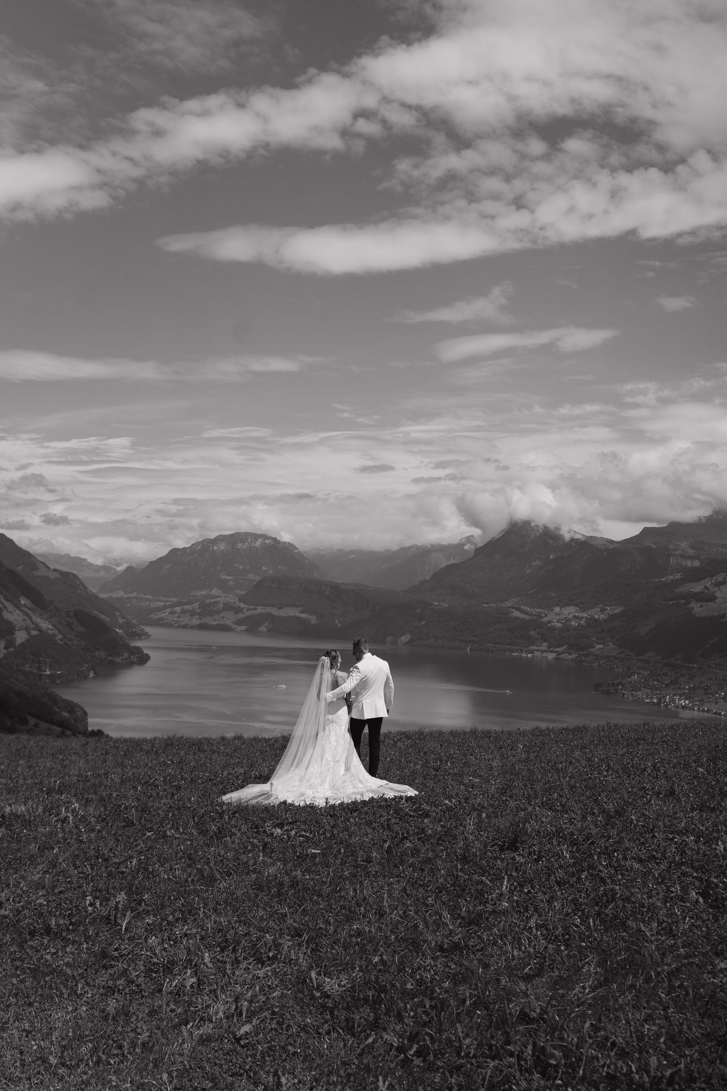Black and white photo of bride and groom standing on a hillside overlooking a stunning alpine lake.