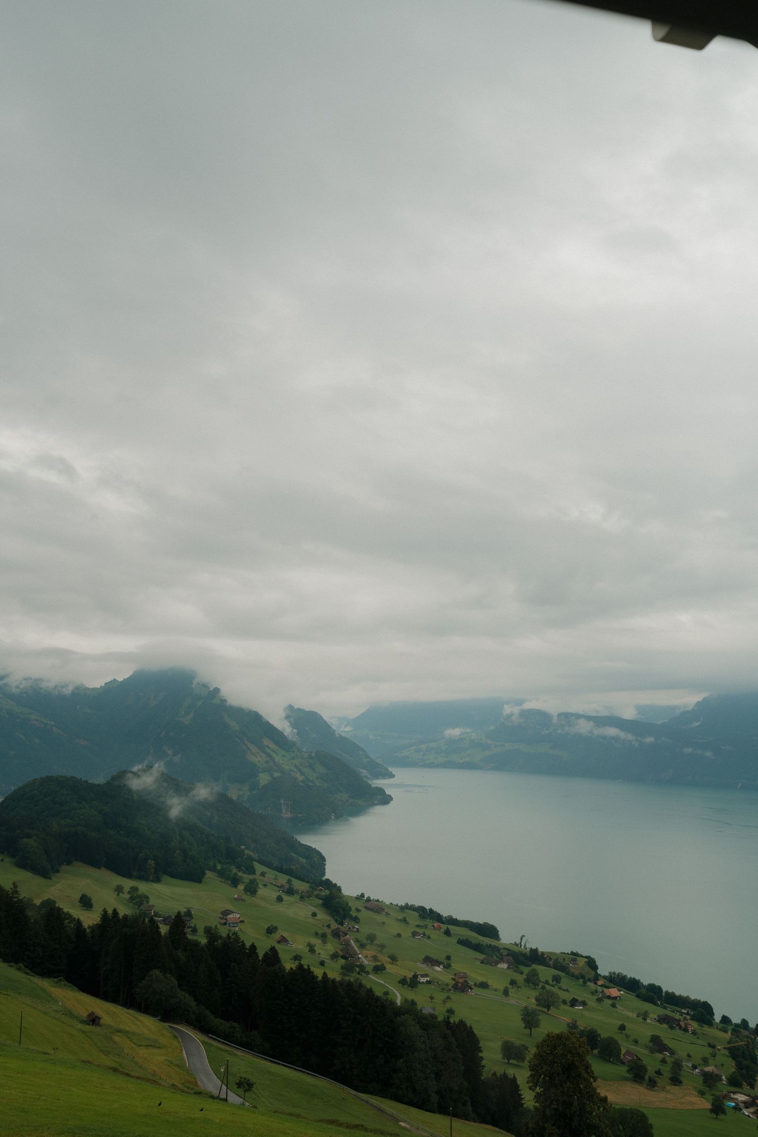 Aerial view of Swiss alpine lake surrounded by green hills and mountains under cloudy sky.