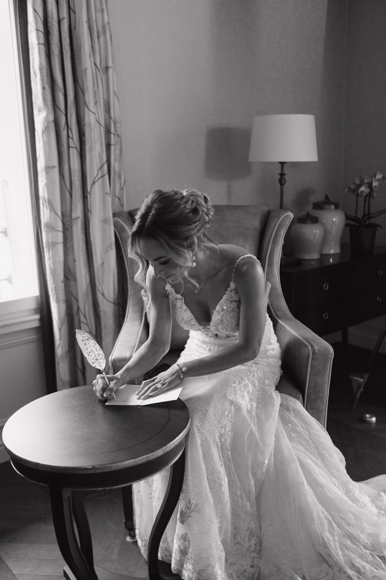 Black and white photo of bride in lace gown writing vows at small table in elegant room.