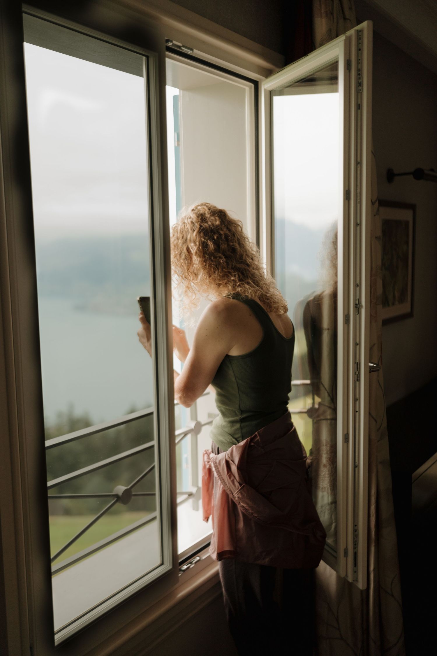 Curly-haired woman standing at open window overlooking a misty alpine lake holding a coffee cup.
